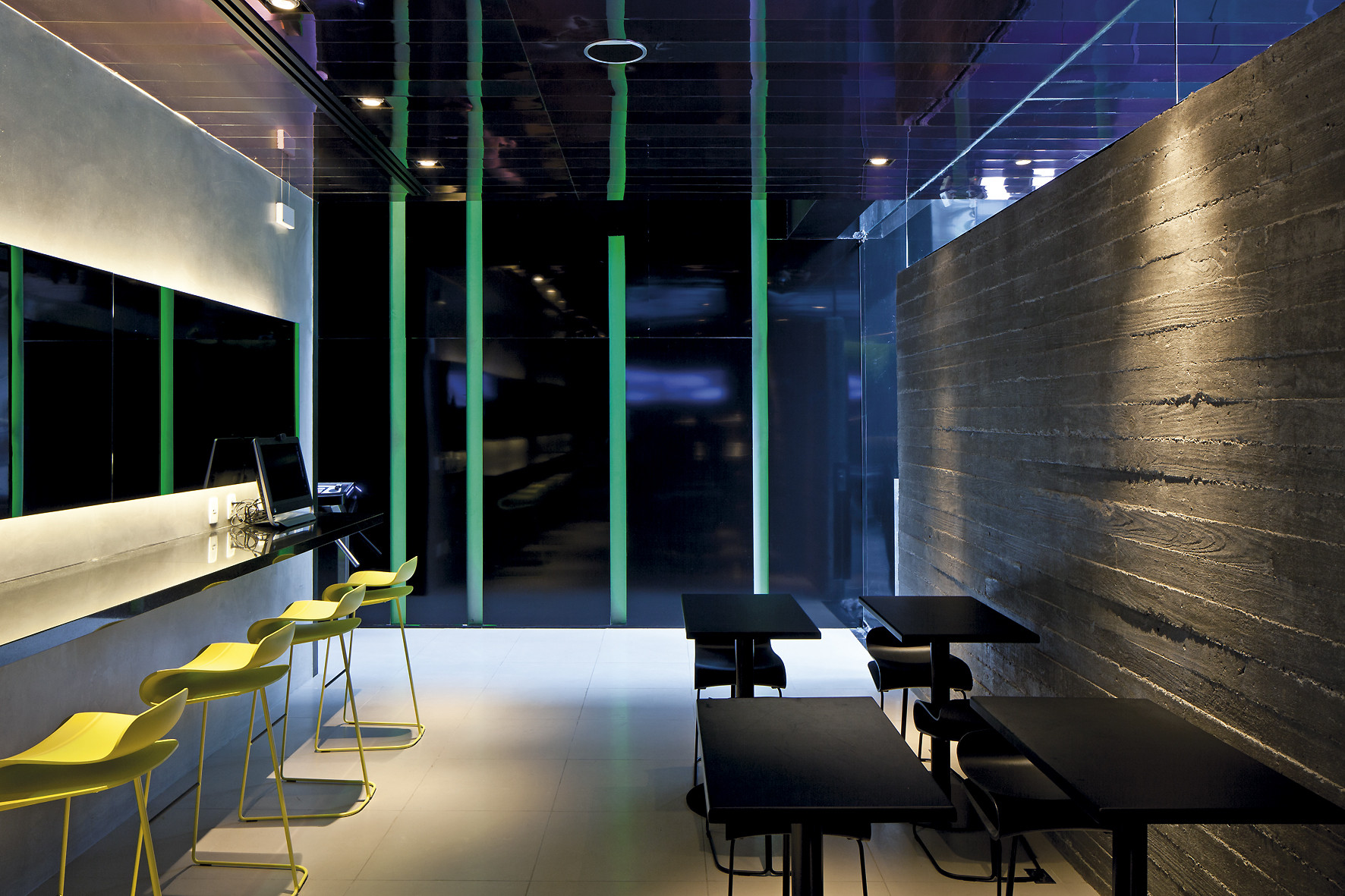 Cafe seating area with yellow stools at a lit counter and black tables beside a board-formed concrete wall at night