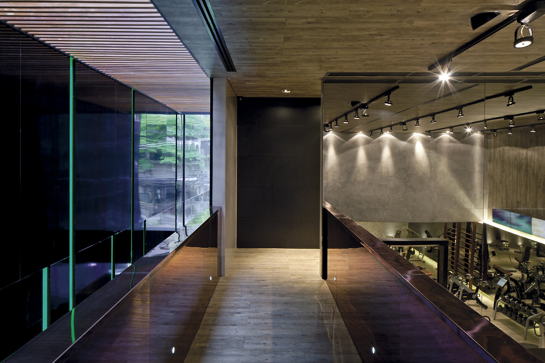 Timber-floored walkway with glass balustrades, looking toward a concrete wall lit by track spotlights above the gym floor