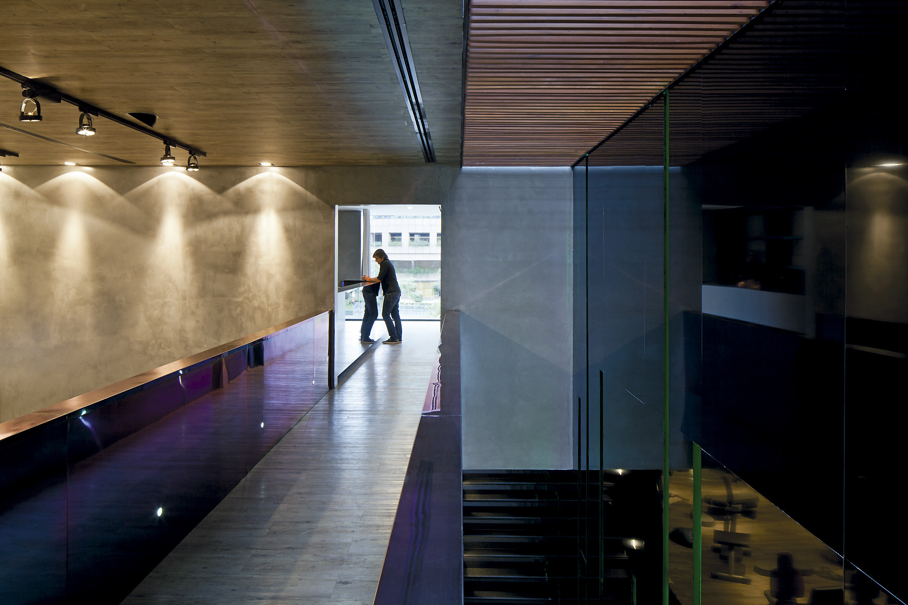 Person standing at the far end of a wood-floored corridor beside a concrete wall washed by ceiling spotlights