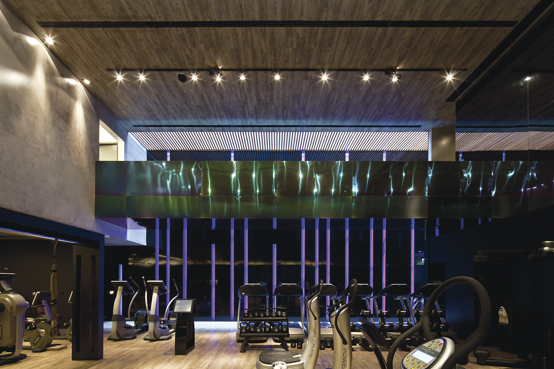 Gym floor with cross trainers and weight racks beneath a timber ceiling and a rippled metal band, with purple light behind