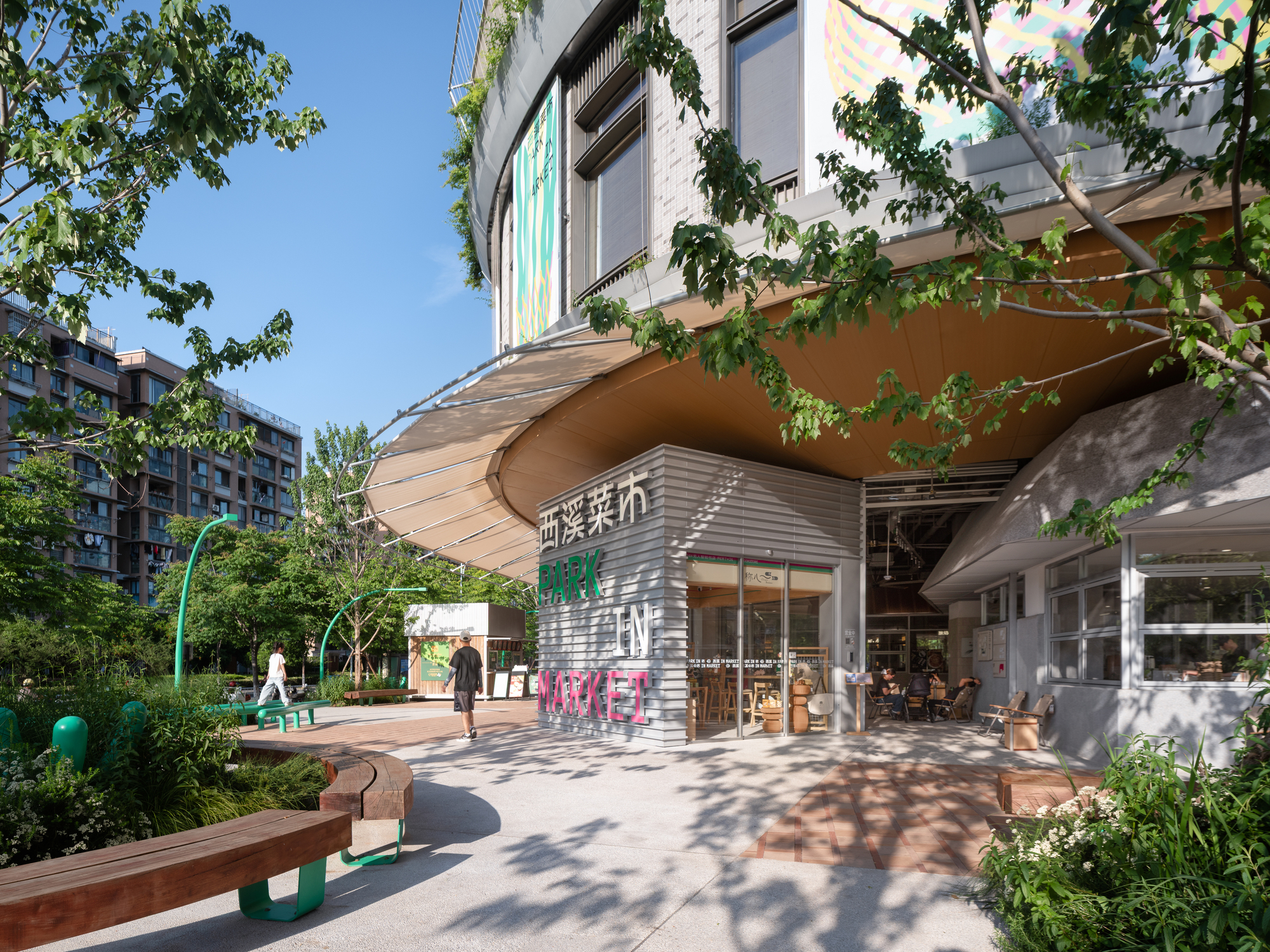 Curved timber-soffit canopy over a market entrance with signage on a grey cladding box, green benches and dappled tree shade
