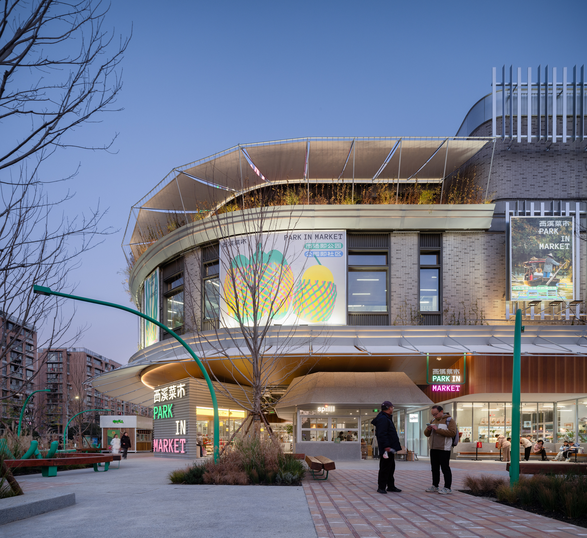 Rounded brick market building with a planted roof edge and large banners, glowing at dusk above people on a paved plaza