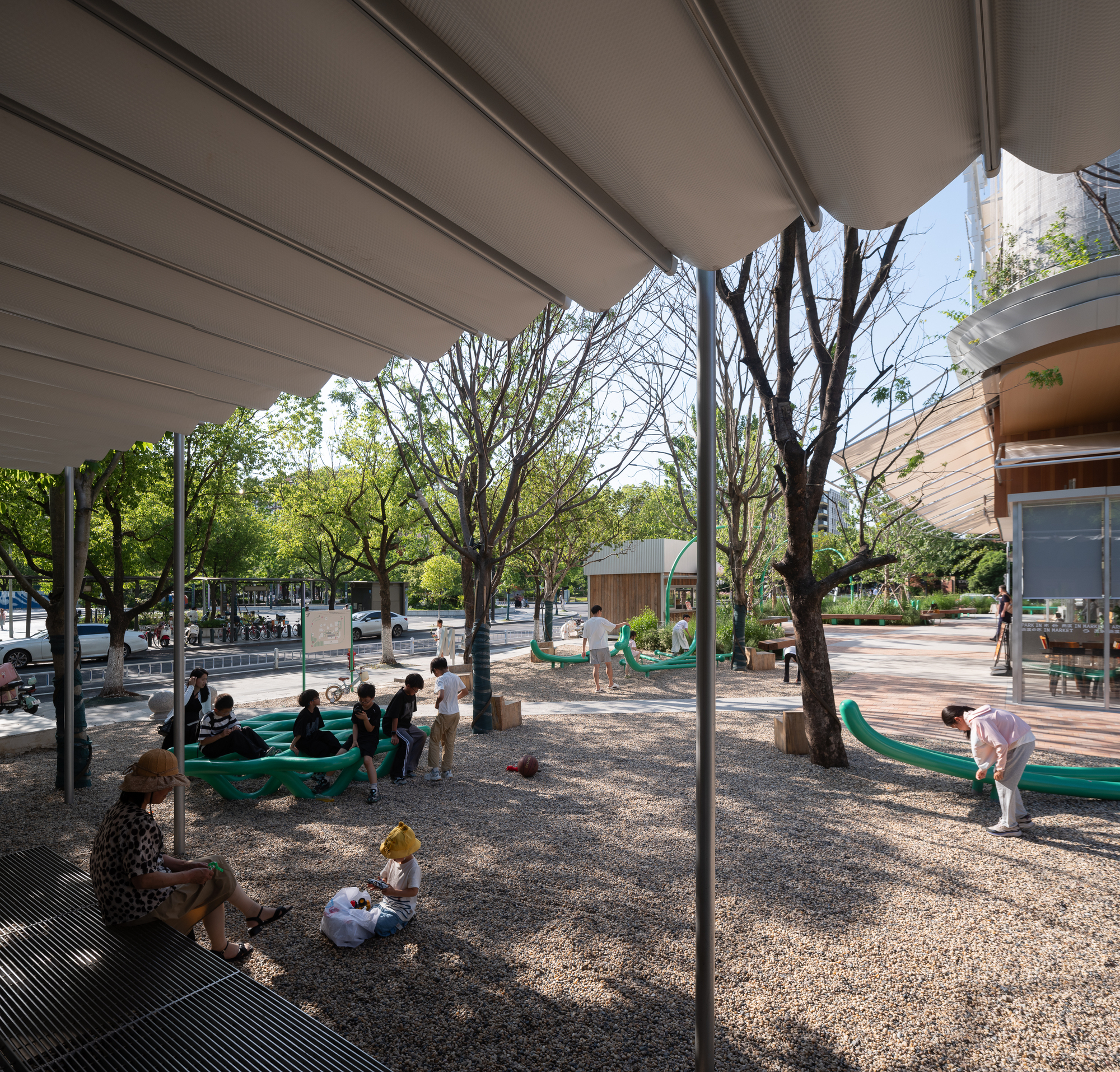 View from beneath a fabric canopy onto a gravel court where children play near green tubular benches and bare trees in sunlight