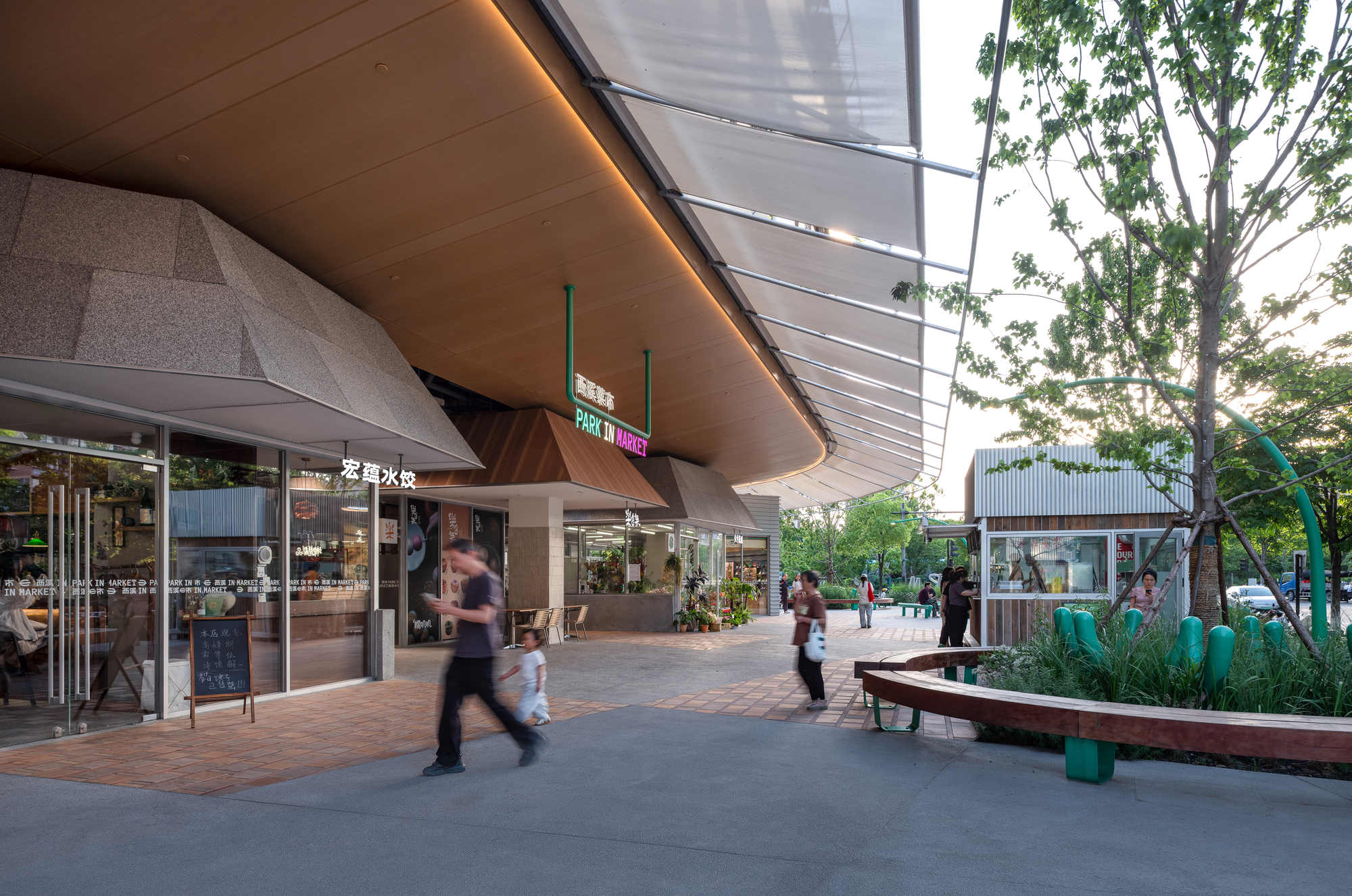 Curved timber soffit and tensioned canopy over shopfronts, with a pedestrian and child walking past at dusk