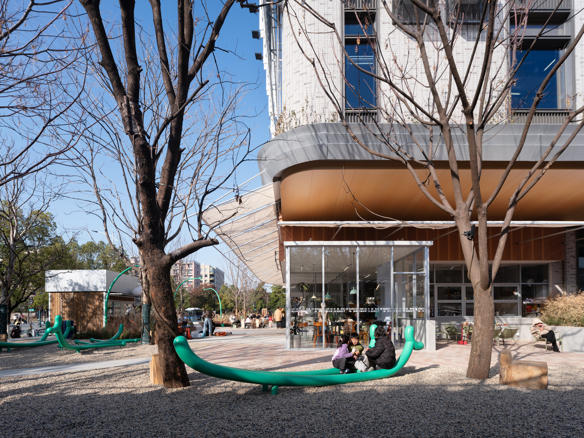 Green looping seesaw in a gravel play area with children, bare winter trees and a glazed cafe behind