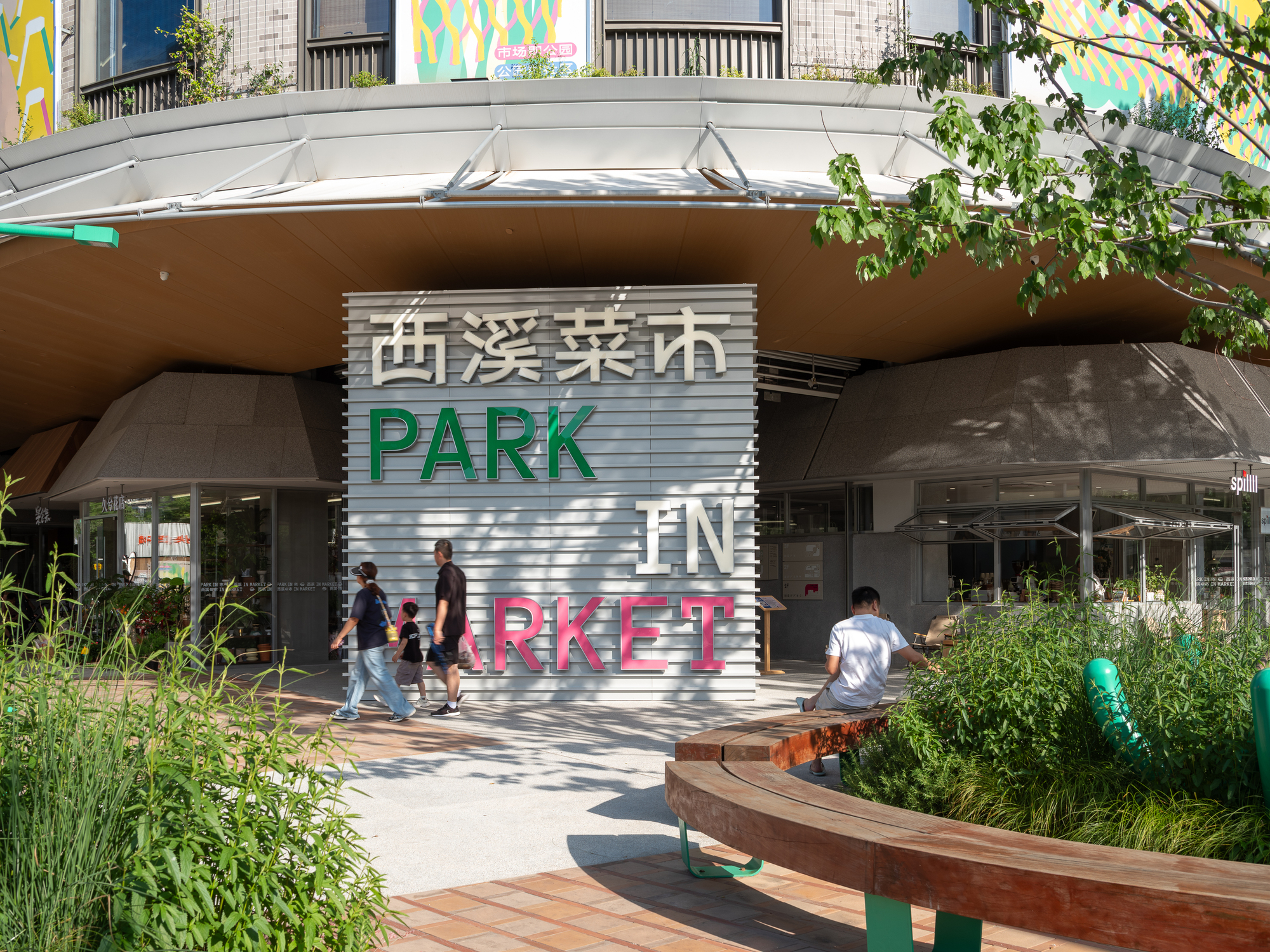 Louvred entrance sign wall with green and pink lettering, a curved timber bench and tall grasses in afternoon sun