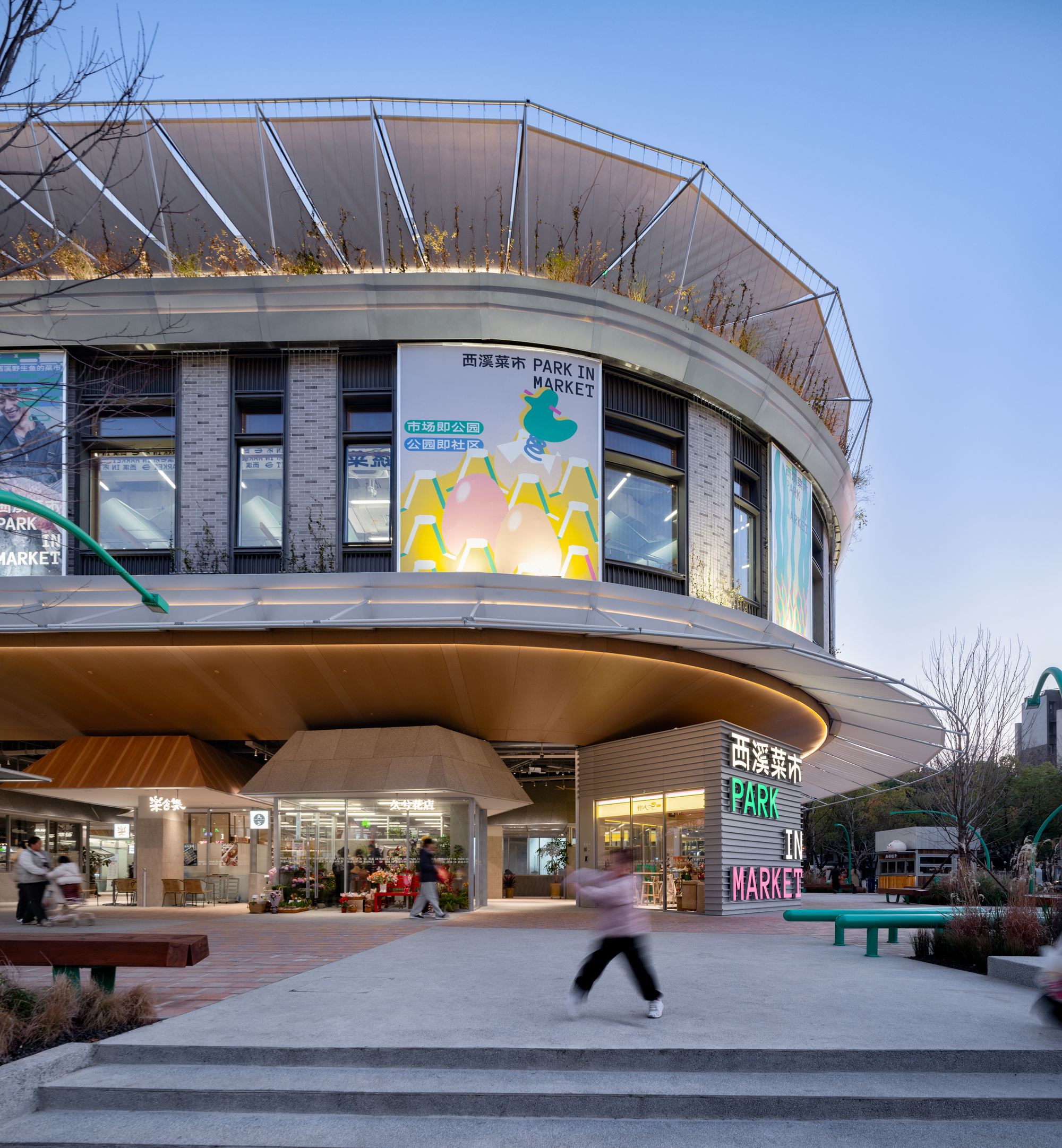 Round market building at blue hour with a planted roof rim, glowing soffit and a blurred child running across the plaza