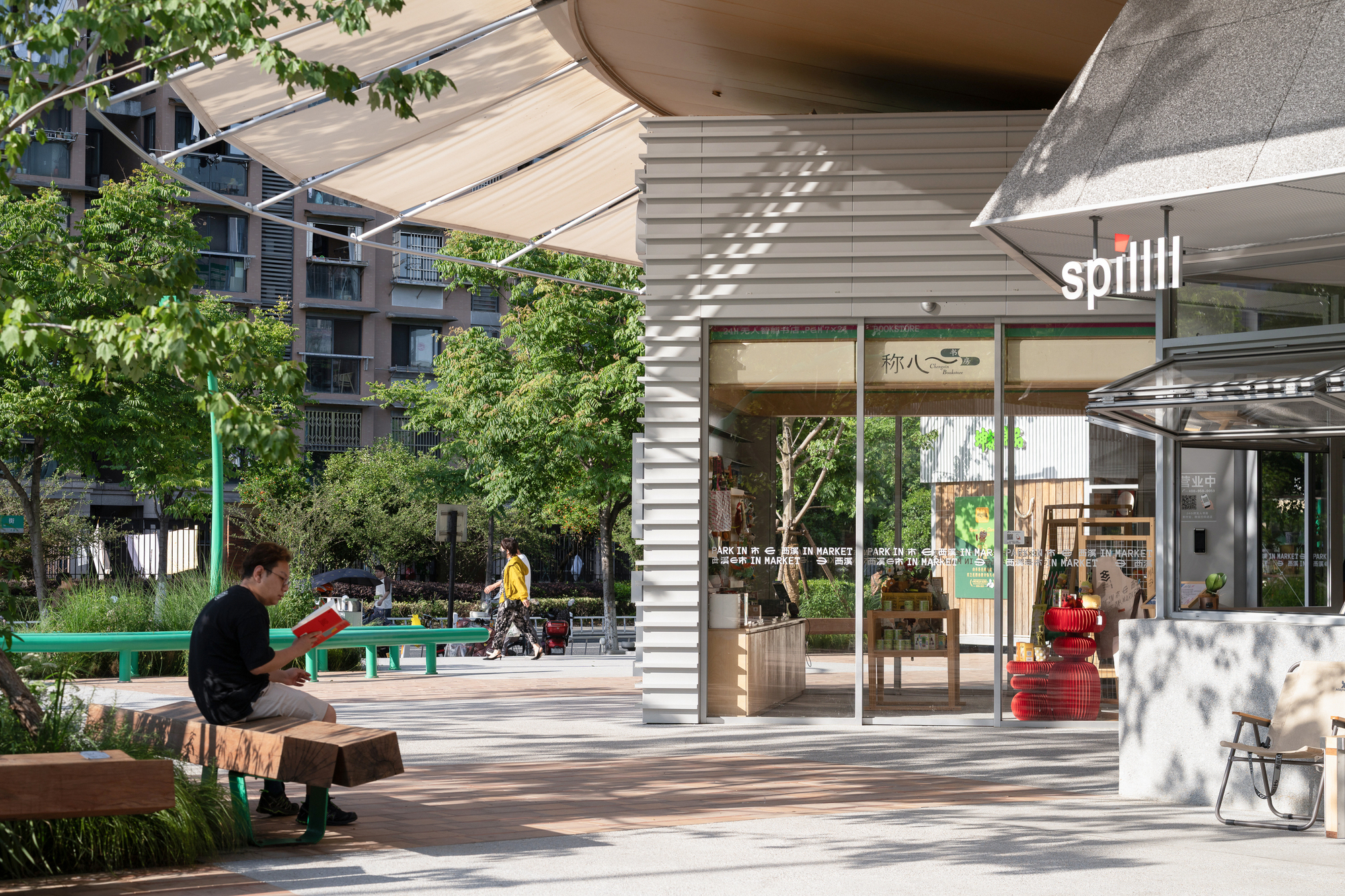 Corrugated metal clad market entrance with glazed front and fabric awning, a man reading on a timber bench in dappled tree shade