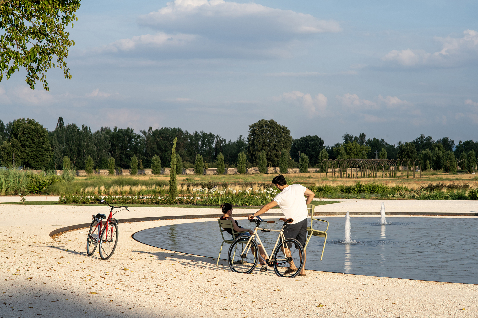 Two cyclists with bicycles beside a shallow circular pool with small fountains, under late afternoon light and scattered clouds