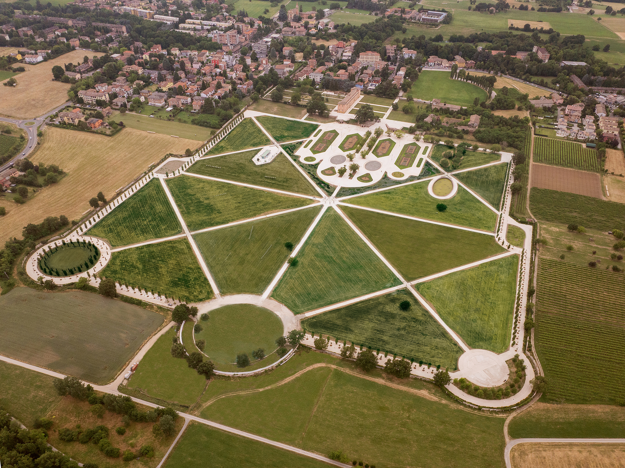 Aerial view of a radiating park plan with wedge-shaped green lawns, circular pavilions and a village beyond, in hazy summer light