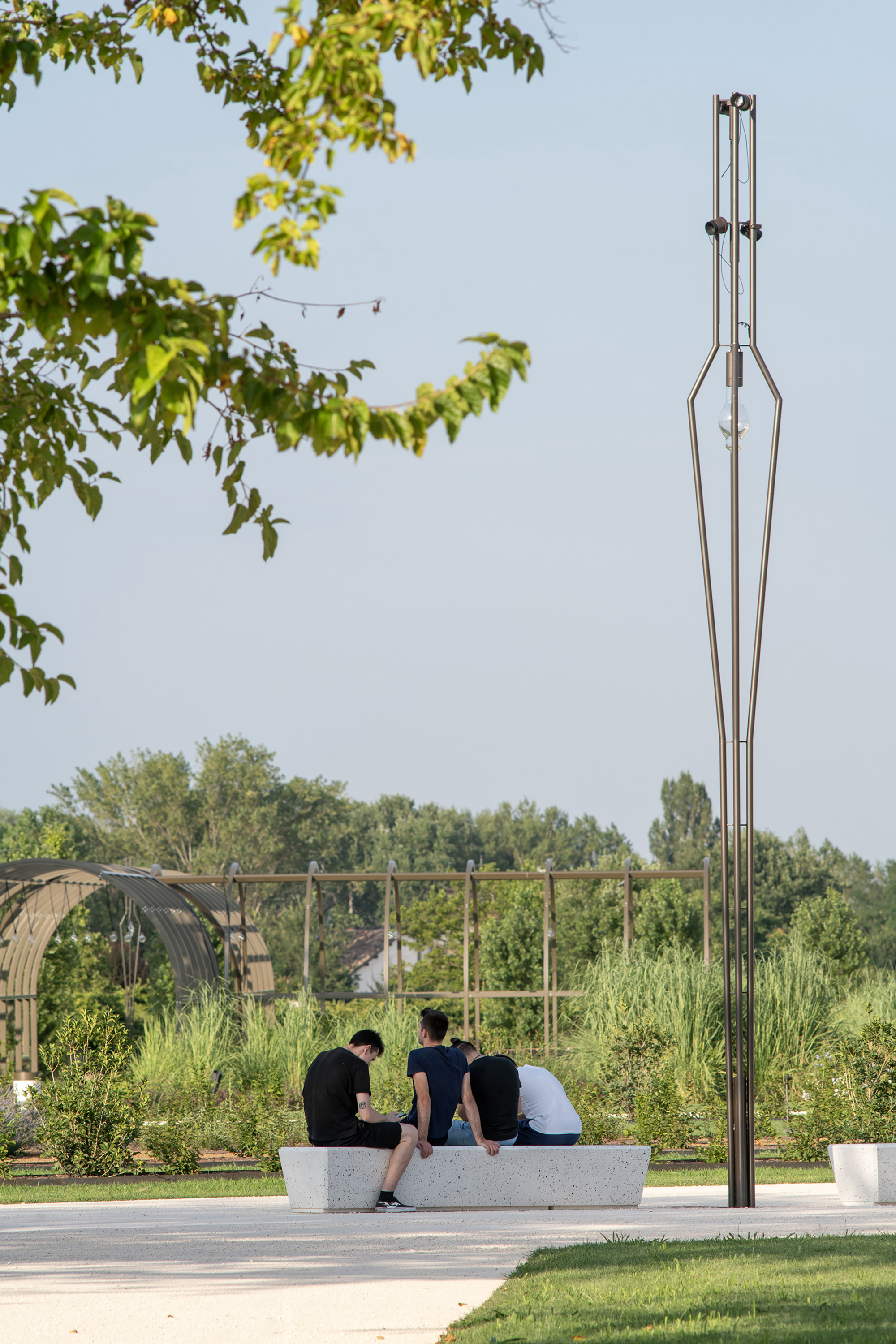 Group of young people seated on a long concrete bench beside a slim metal lamp post, with a pergola behind