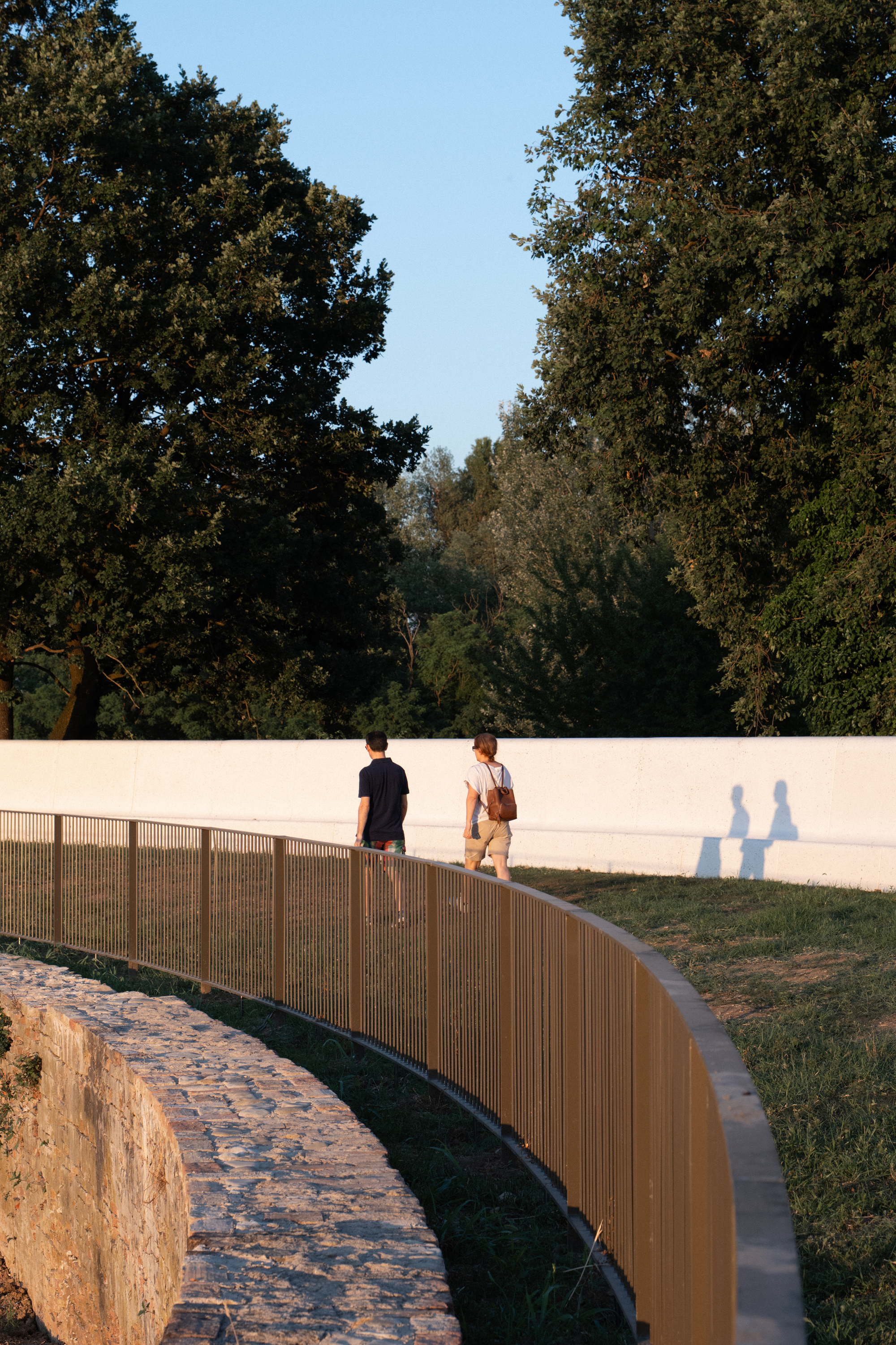 Curved golden metal railing above a rough stone retaining edge, with two walkers passing a white wall at sunset
