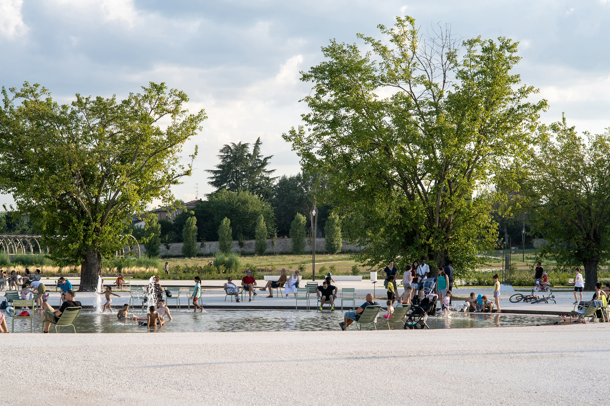 Families and children gathered around a shallow fountain pool with green metal chairs, beneath large trees on a cloudy summer afternoon