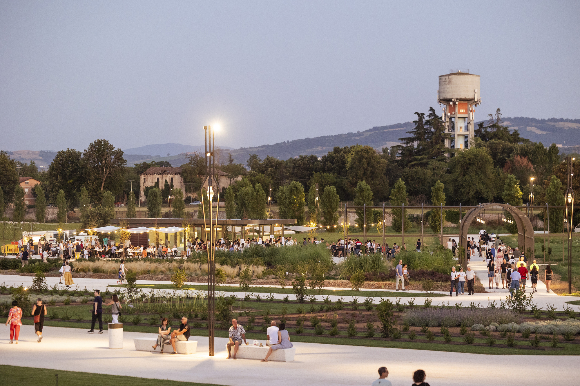 Visitors seated on white benches along a lit garden path at dusk, with a water tower on the hill behind