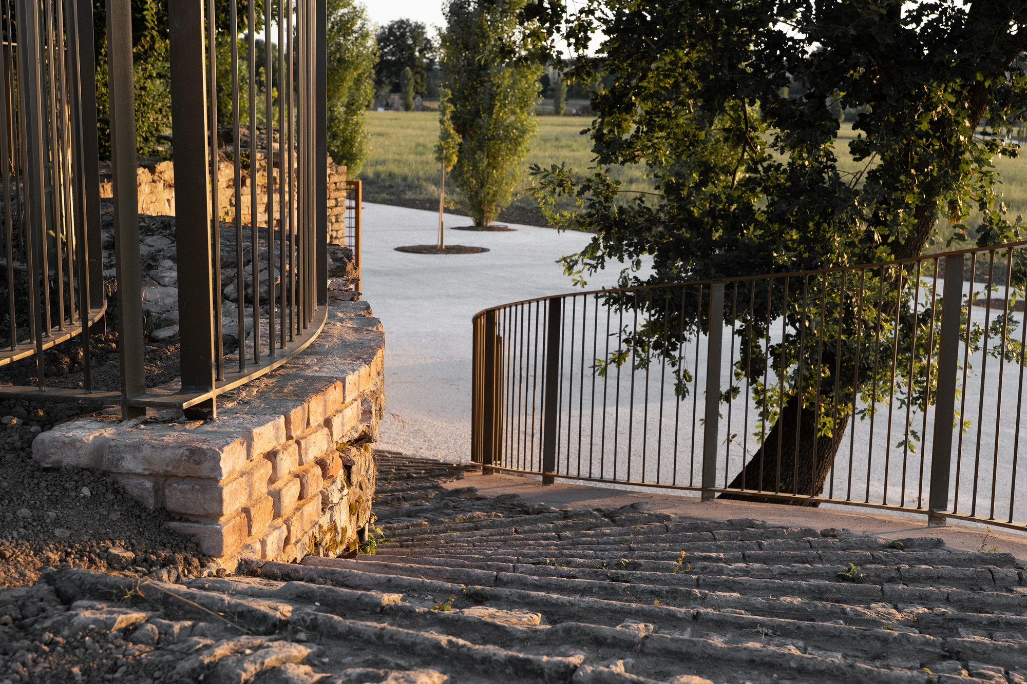 Worn stone steps beside a curved brick retaining wall and steel railing, lit by low evening sun through leaves