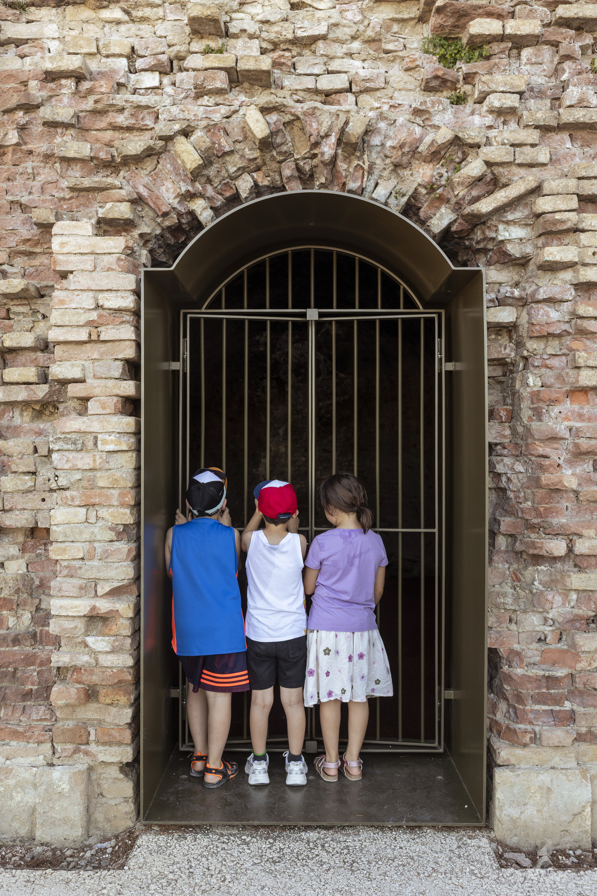 Three children peering through a barred gate set in an arched opening of weathered brick wall