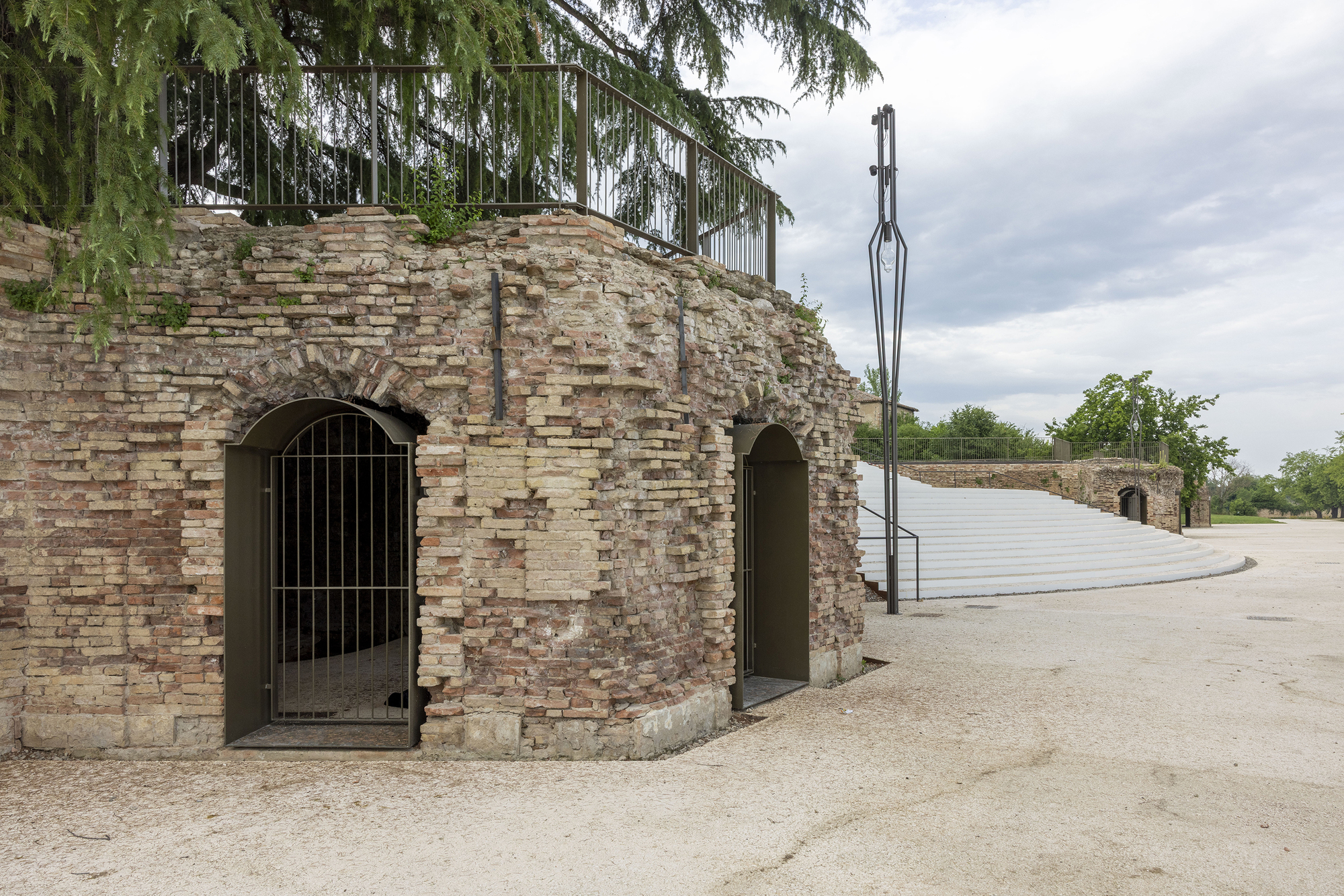 Old brick bastion with arched gated openings and a railing above, beside a white stepped amphitheatre under overcast skies