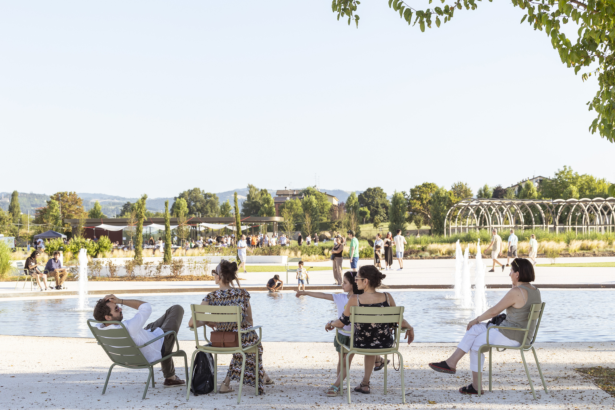 Visitors seated on green metal chairs beside a shallow pool with fountain jets in warm late afternoon sun