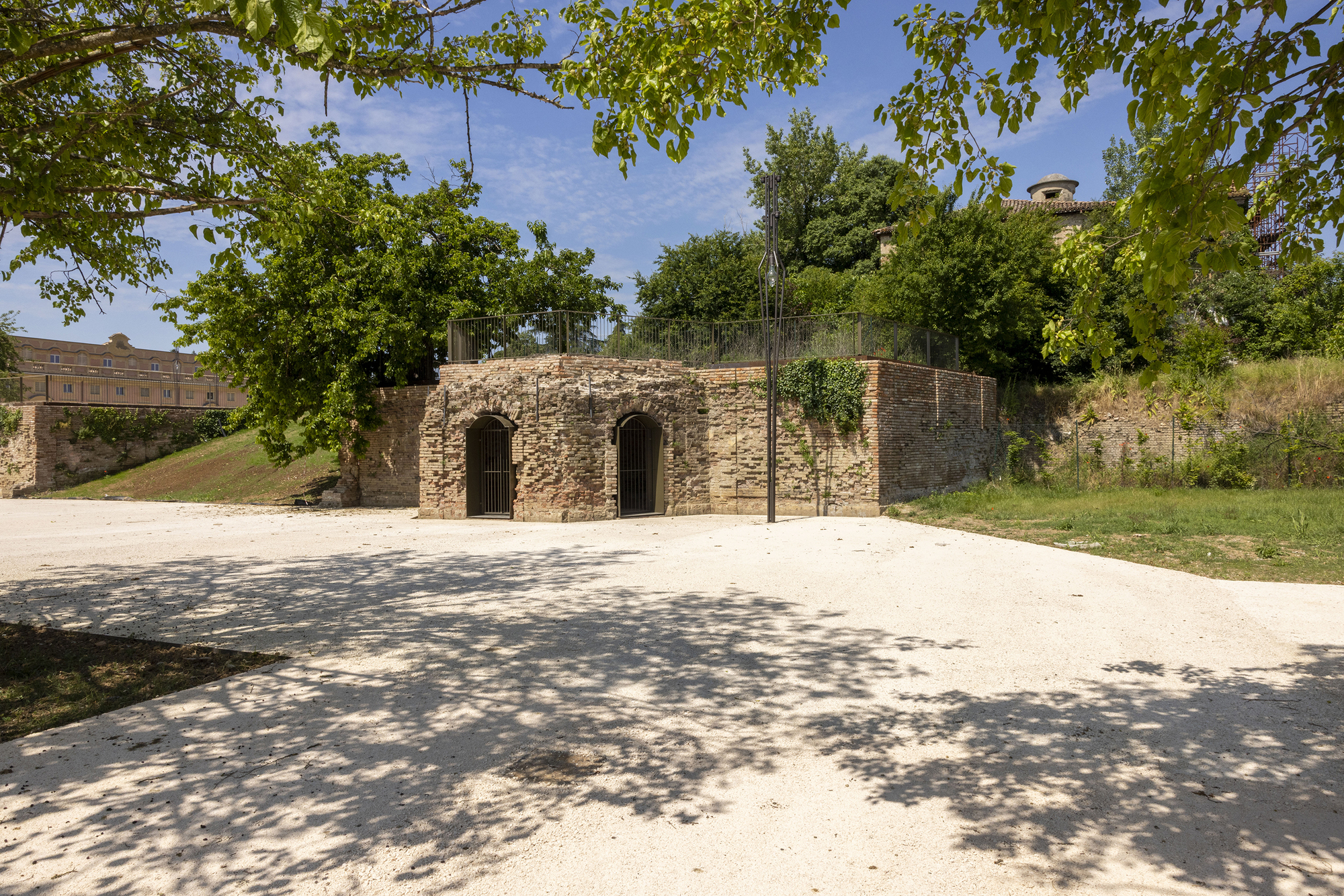 Weathered brick wall with two arched barred openings beside a gravel clearing dappled by tree shadows