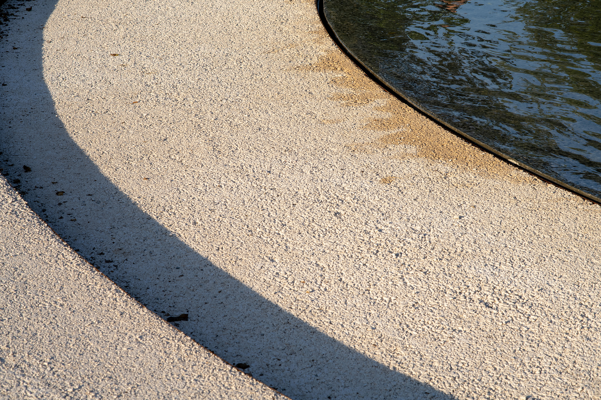 Curved edge of pale gravel meeting dark water, split by a long arc of shadow in low sun