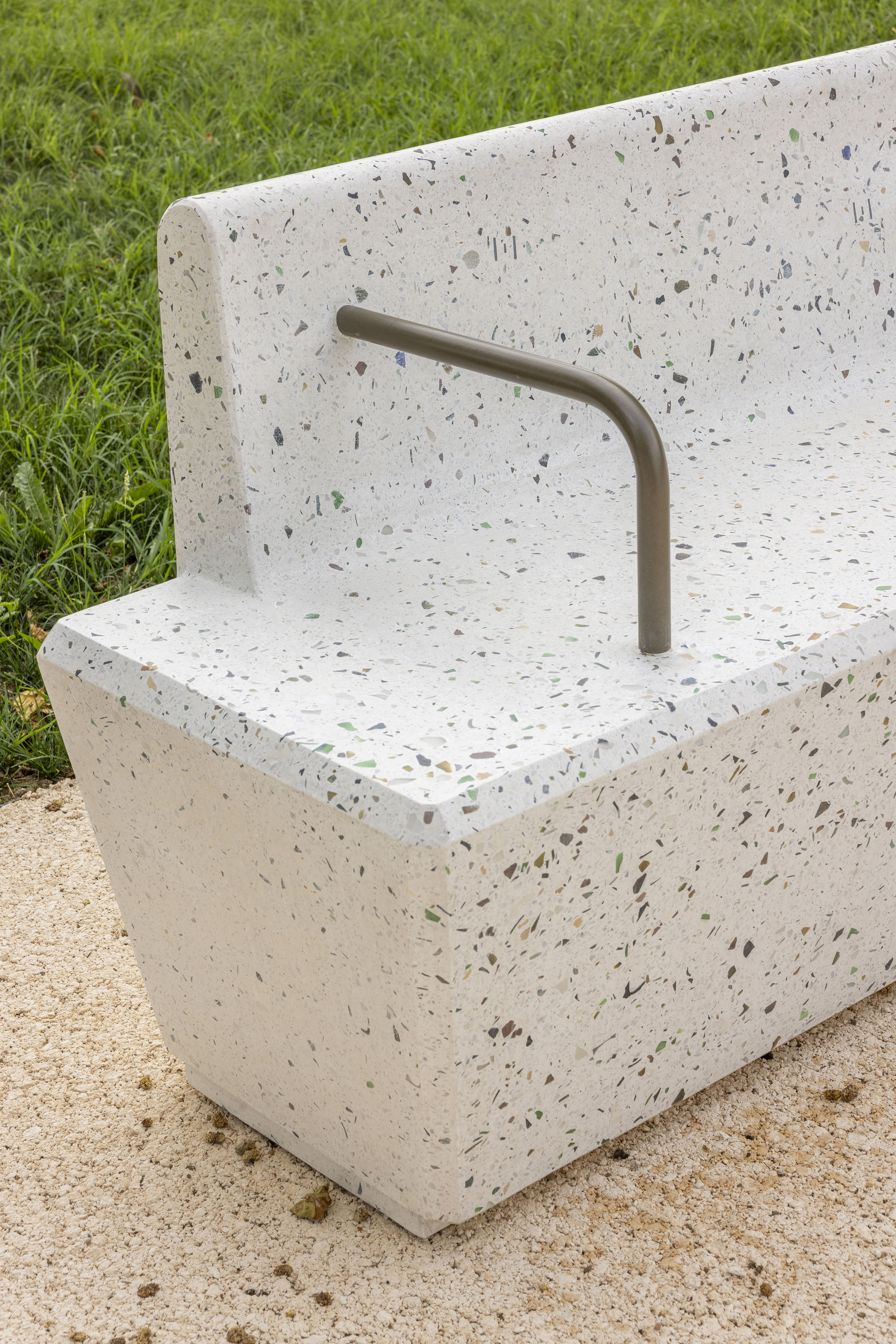 Terrazzo bench with flecked aggregate and a bent metal armrest standing on gravel next to green grass
