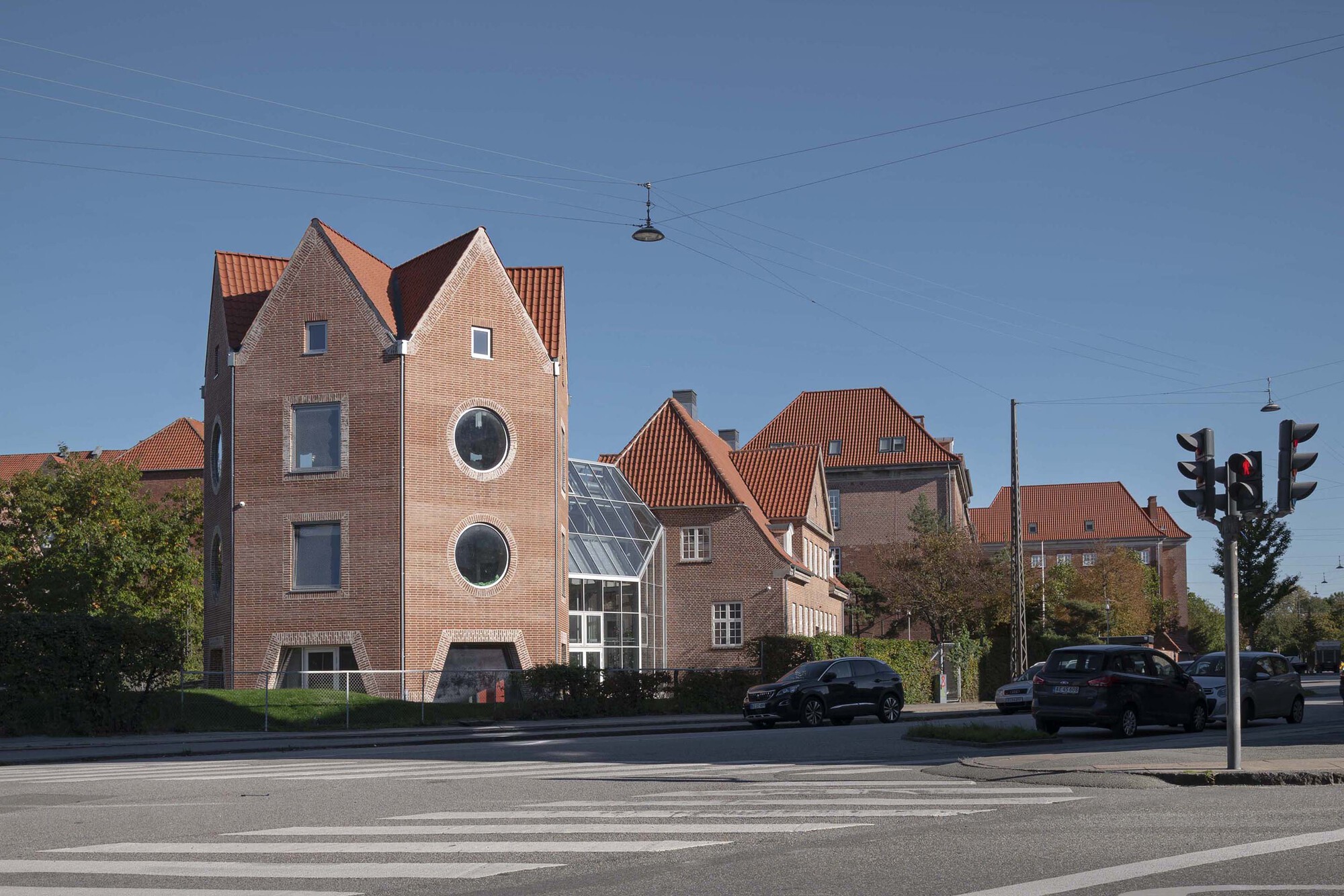 Brick tower with gabled red-tile roofs beside a glazed conservatory, seen across a crosswalk under a clear blue sky
