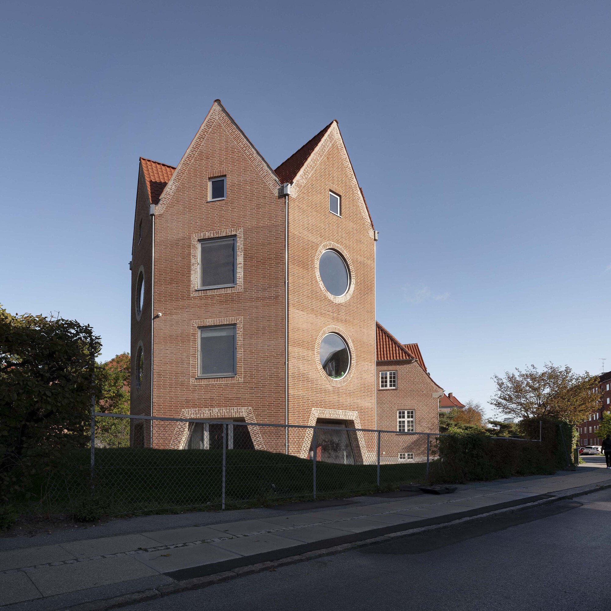 Corner view of the tall brick gables with round windows and arched stone surrounds behind a hedge and fence in low sun