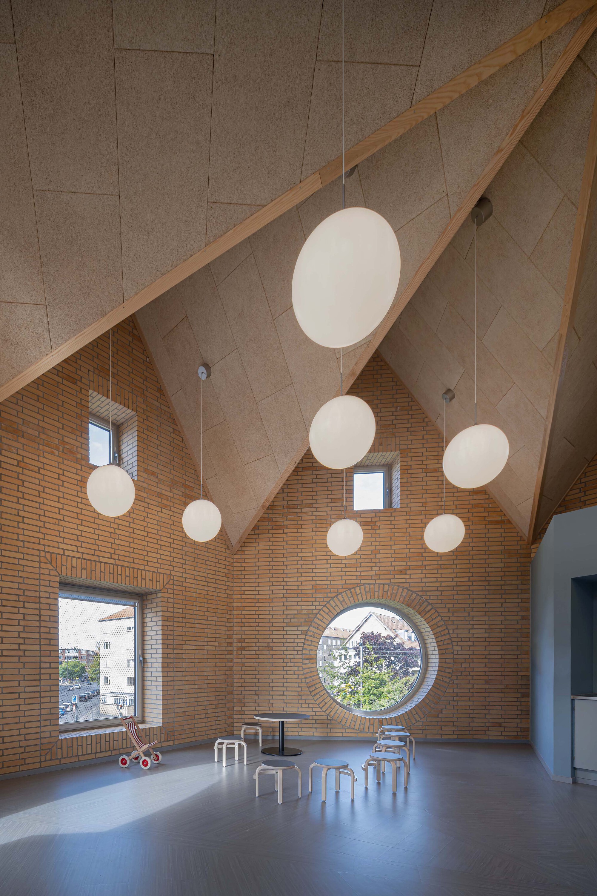 Double-height room with pitched fibreboard ceiling, round window and hanging globe lights, with low stools and a toy pram in sunlight