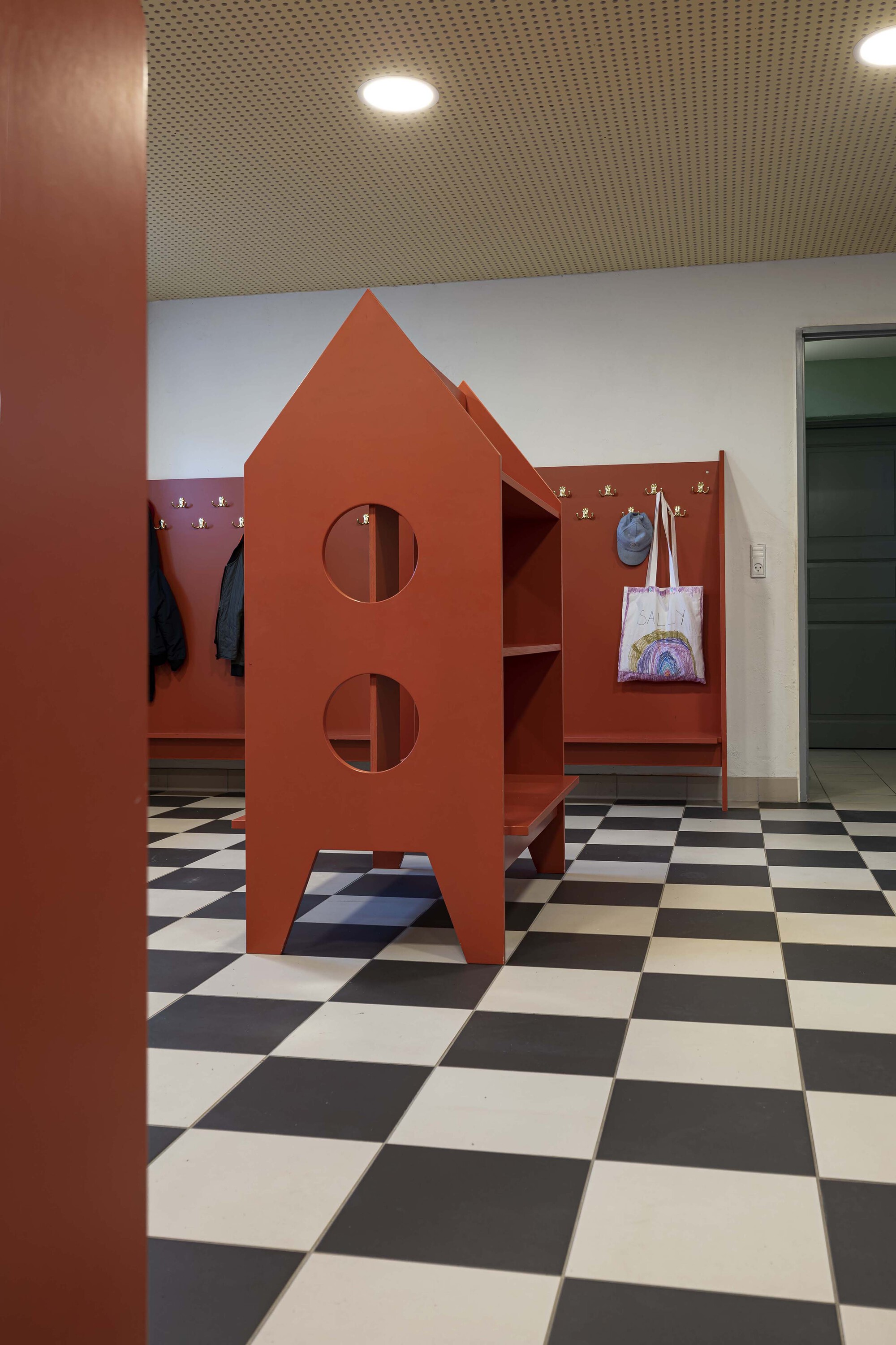Cloakroom with a house-shaped red shelf unit, coat hooks and a hanging tote bag over a black and white checkered floor