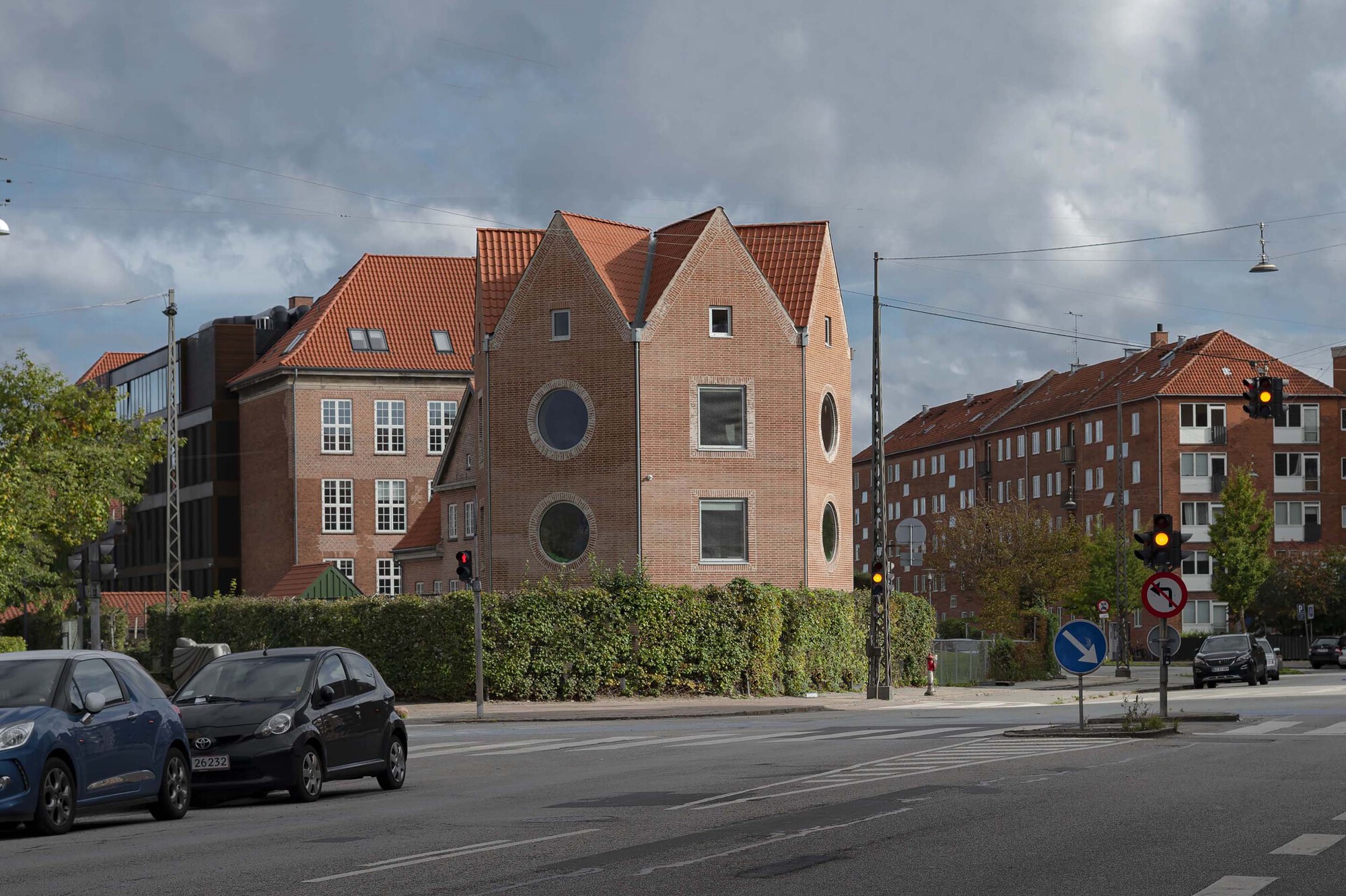 Brick gabled tower with round windows behind a trimmed hedge, seen from a junction with traffic lights under heavy clouds