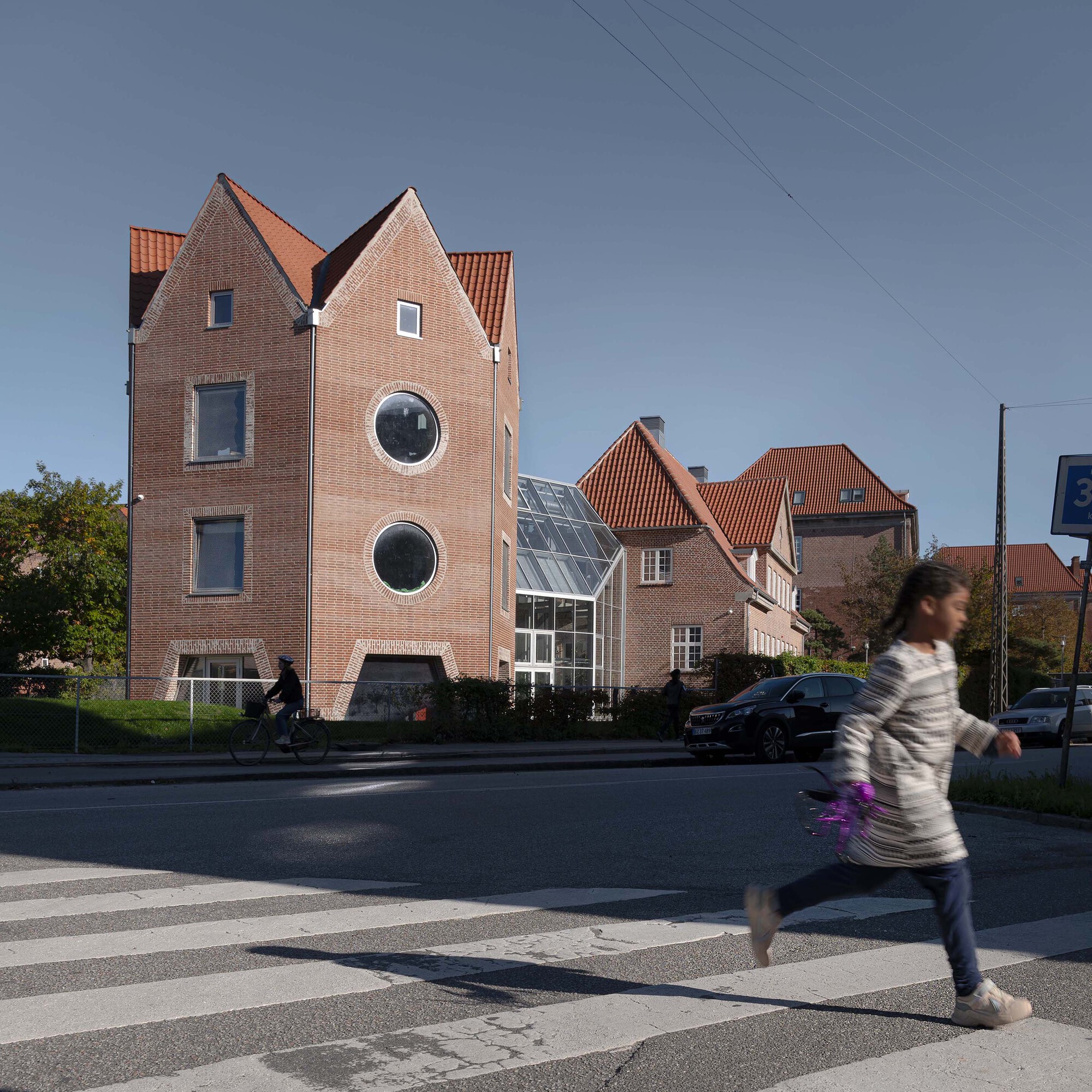 Gabled red brick facade with two round windows beside a glazed link, seen across a crosswalk where a girl runs and a cyclist passes