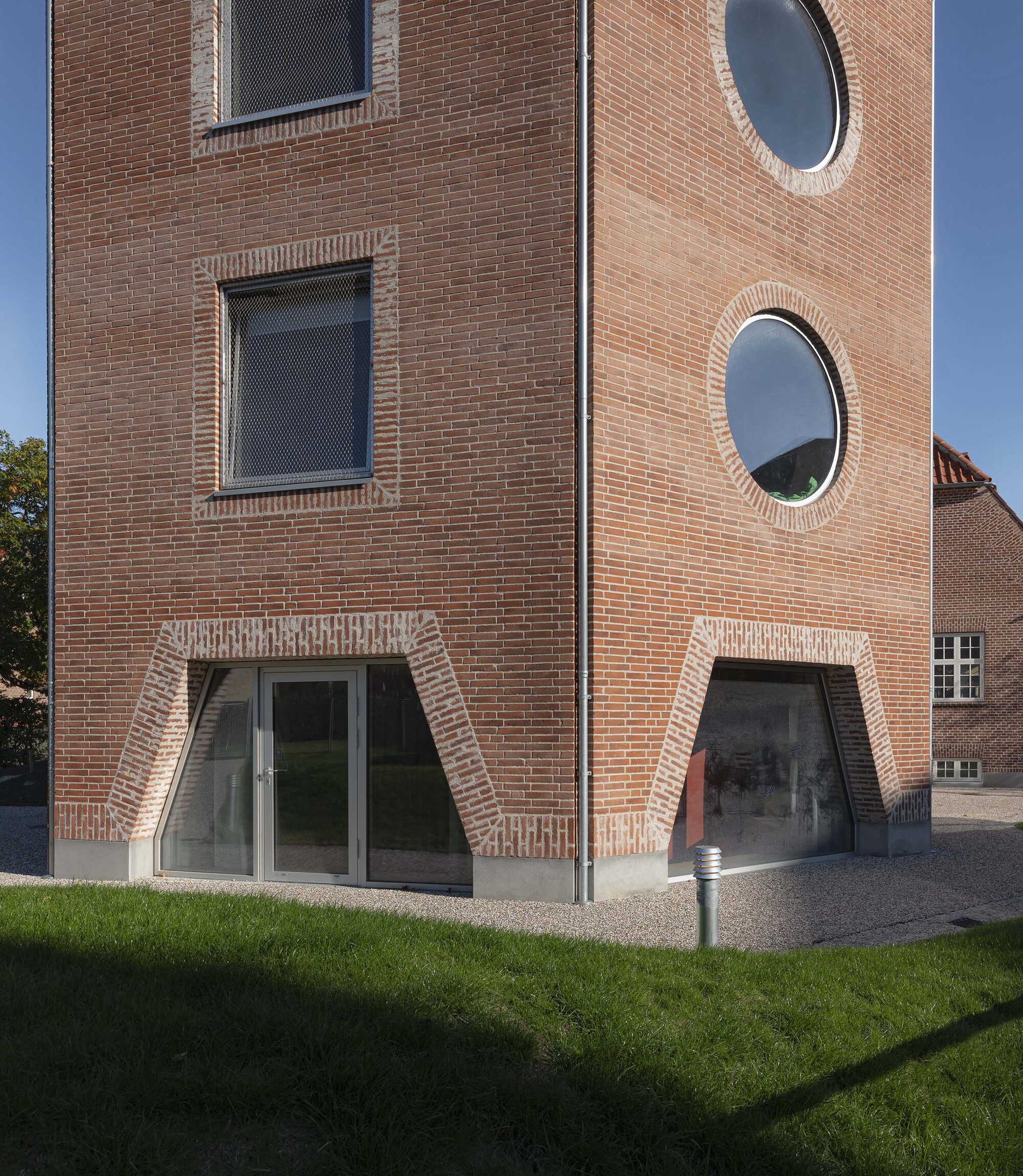 Corner of the brick tower with round windows and angular brick-framed ground floor glazing above a gravel path and lawn in low sun