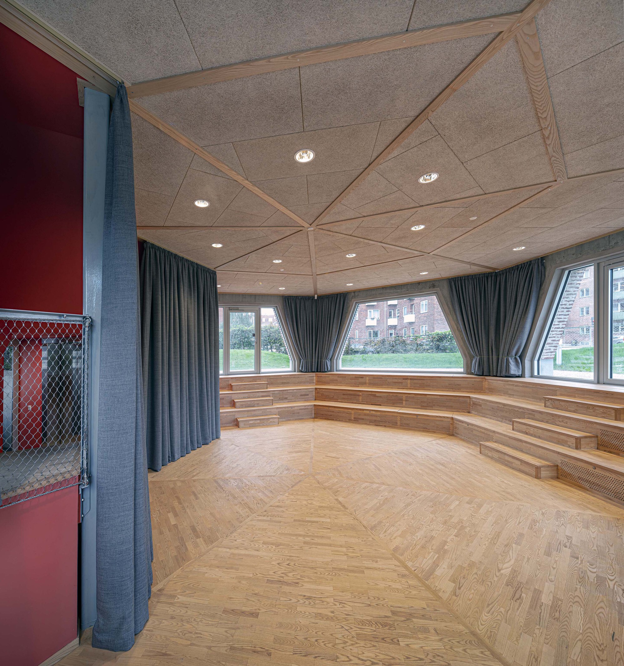 Interior room with tiered timber seating, parquet floor, faceted ceiling panels and grey curtains drawn back from windows facing a green lawn