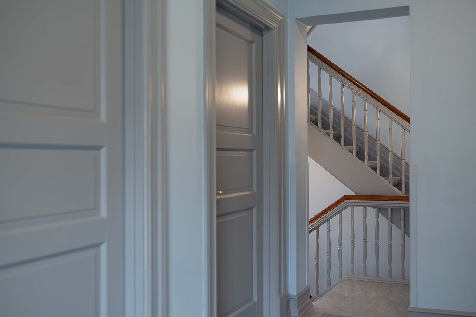 View through a doorway to a painted stair with turned balusters and a timber handrail, framed by panelled grey doors