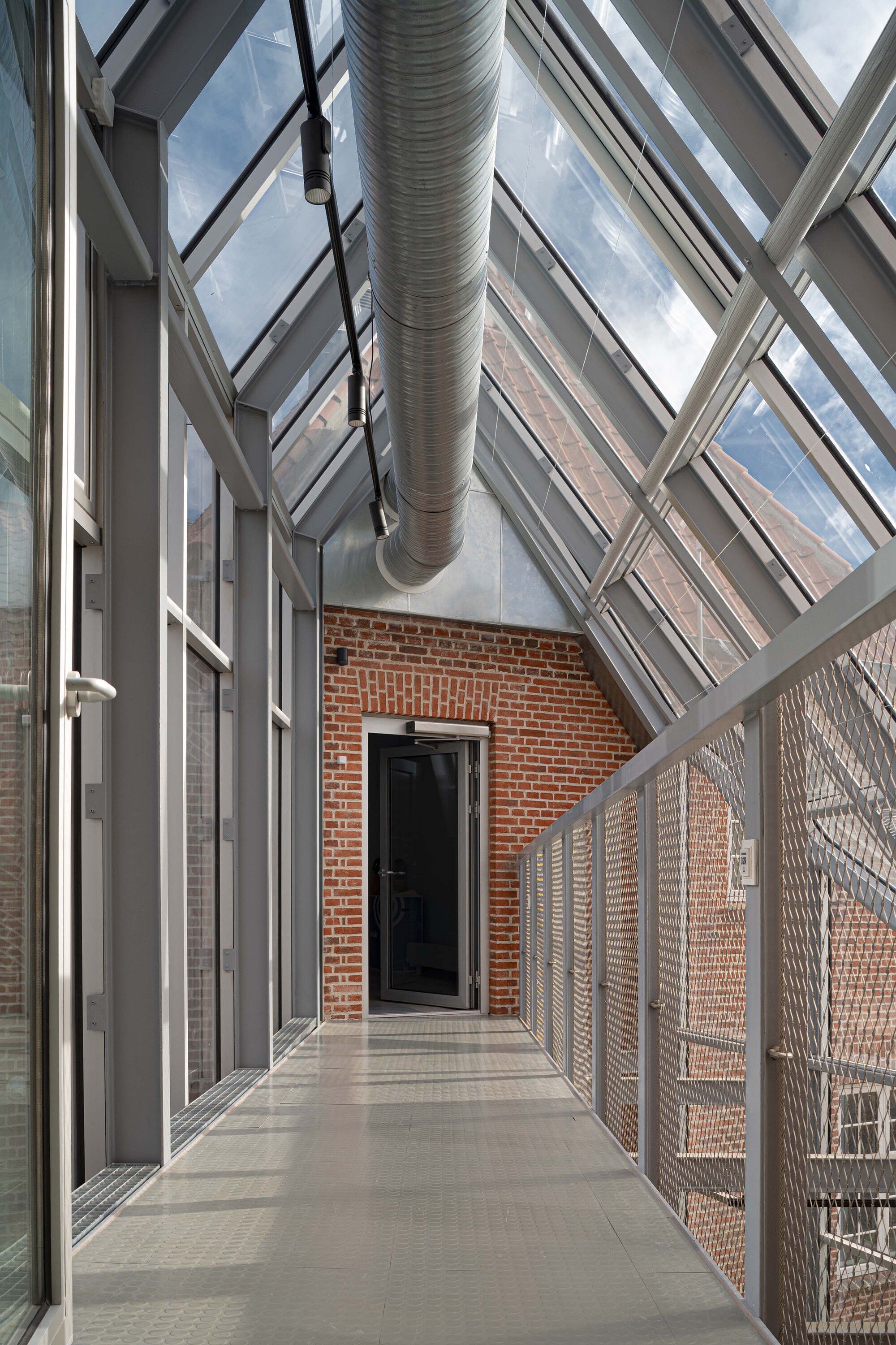 Narrow steel and glass walkway under a pitched glazed roof with a ventilation duct, leading to a brick wall and door