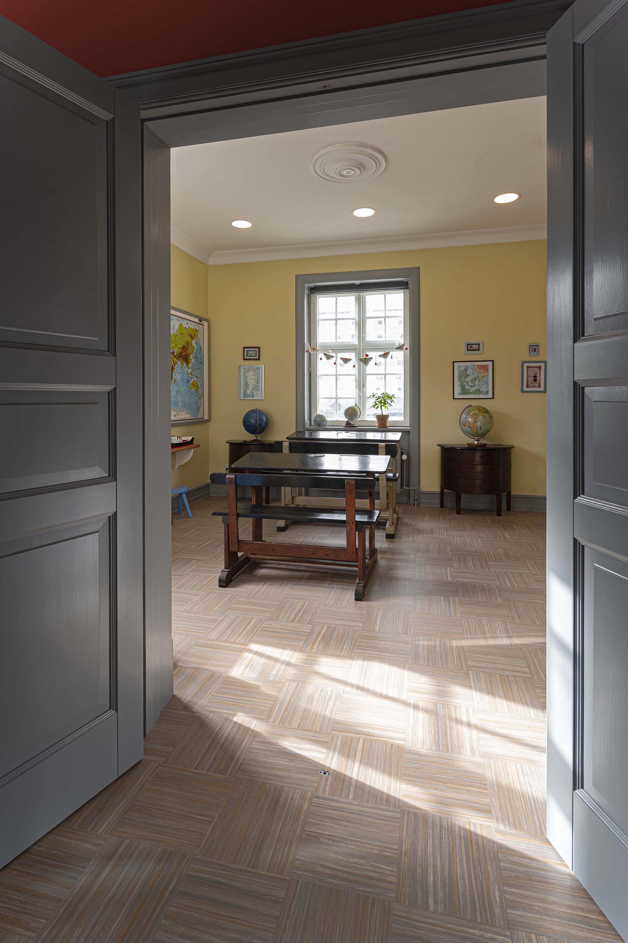 Open grey double doors frame a classroom with wooden school benches, globes and yellow walls, sunlight falling across the parquet floor
