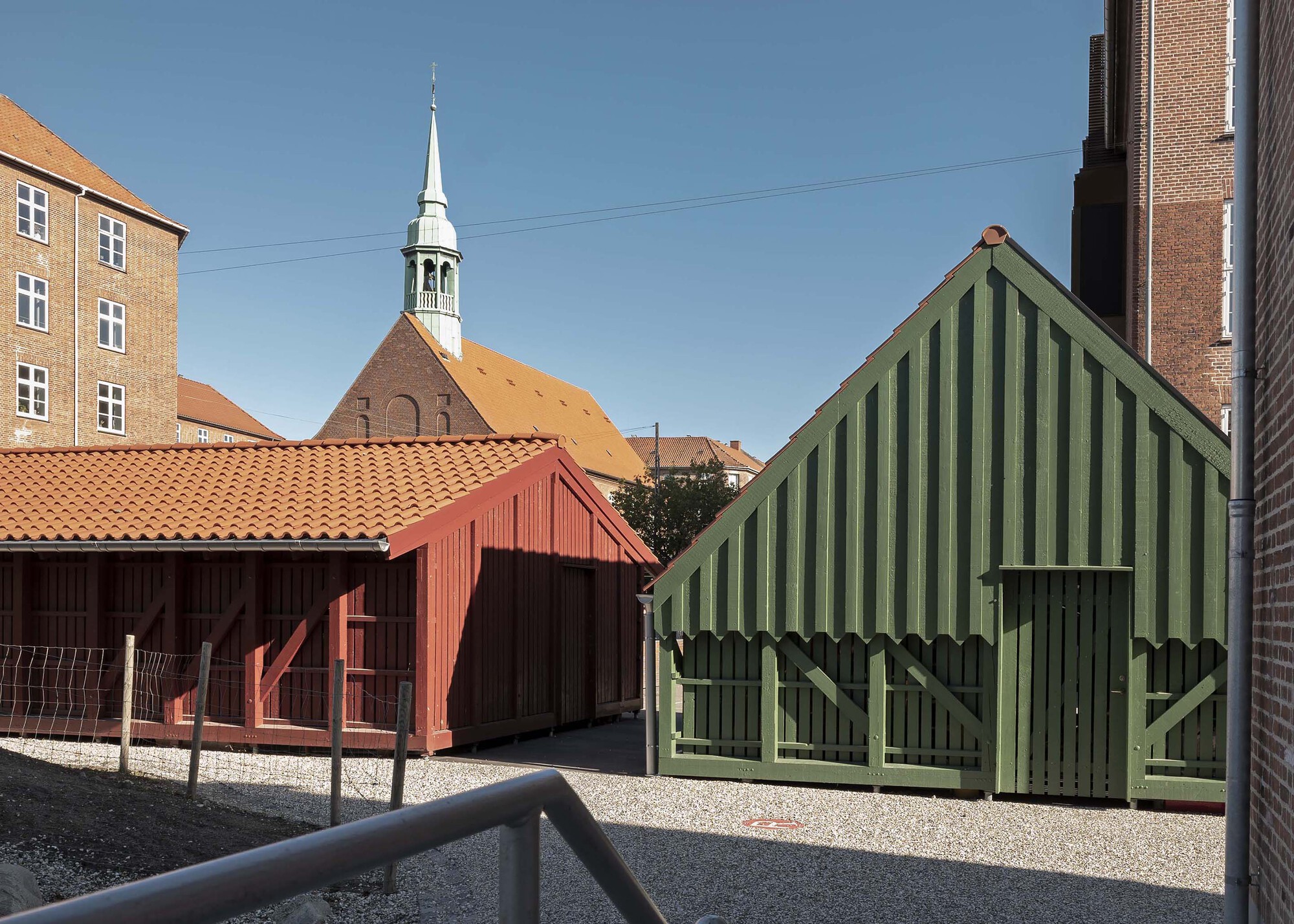 Red and green timber sheds with clay tile roofs on a gravel yard, a copper-green church spire behind under clear sky