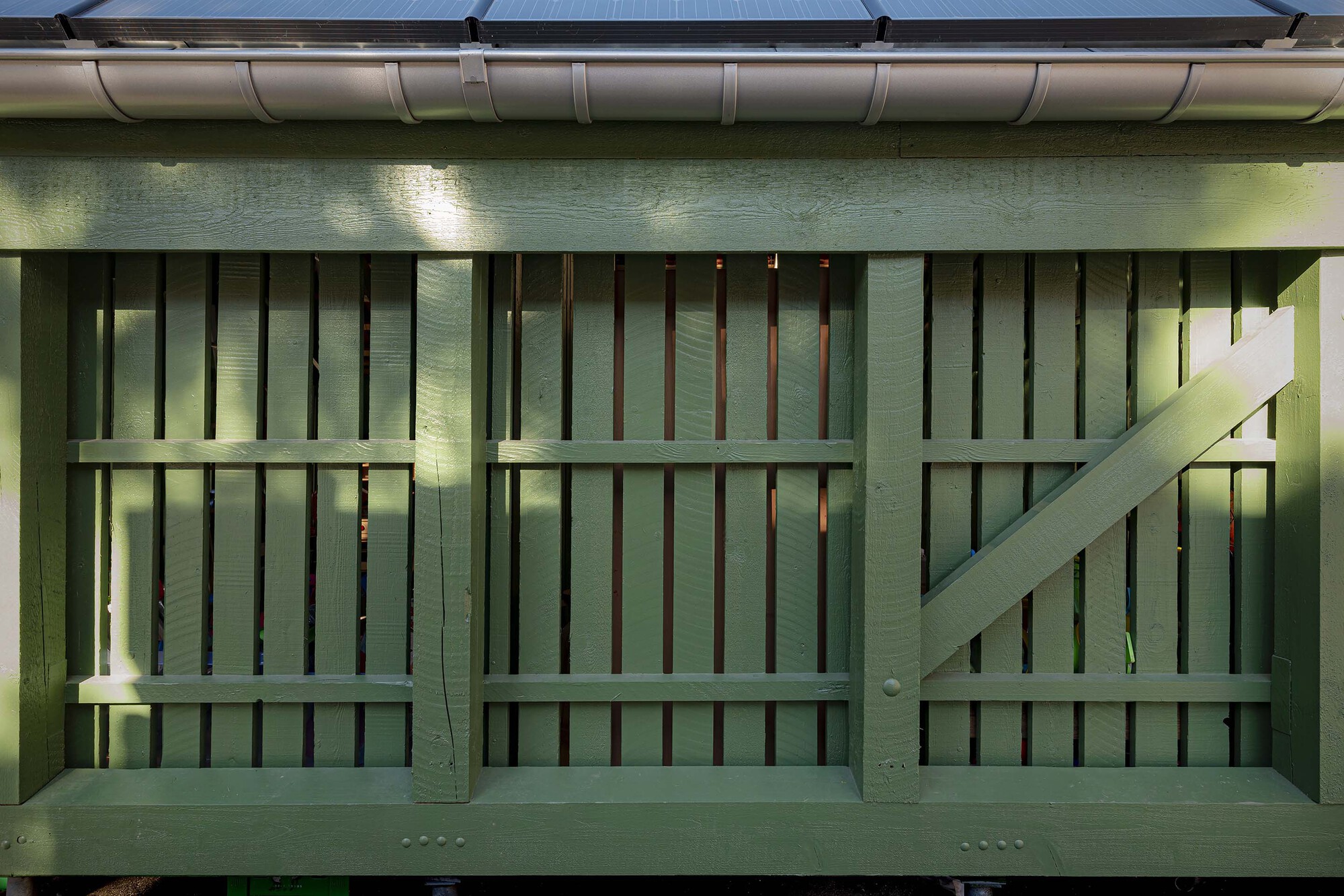 Close-up of the green timber frame with vertical slats and a diagonal brace, dappled sunlight under a gutter and solar panels