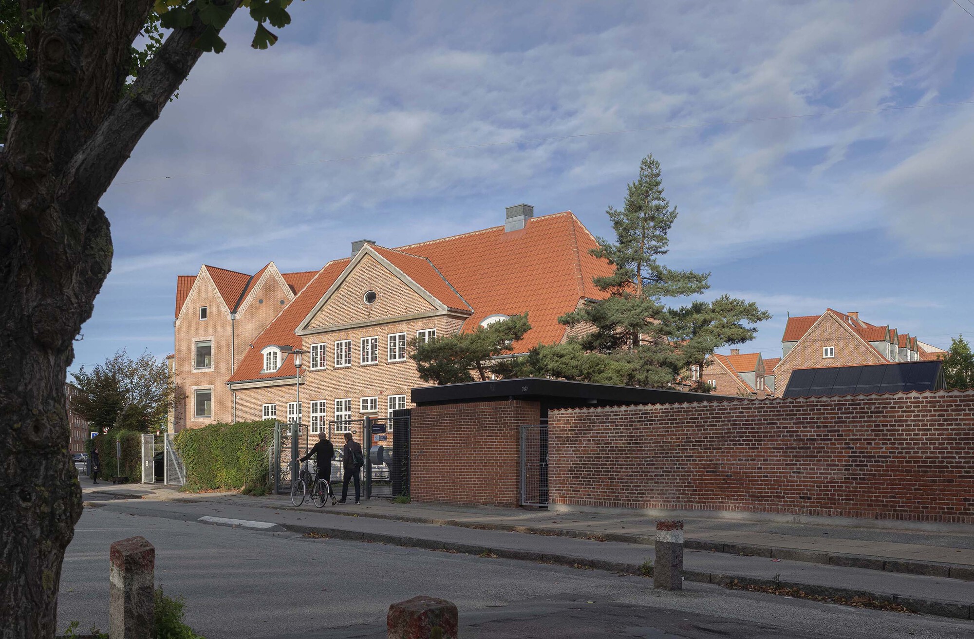 Brick school building with orange tiled roof beyond a low brick wall, two people with a bicycle on the pavement at golden hour