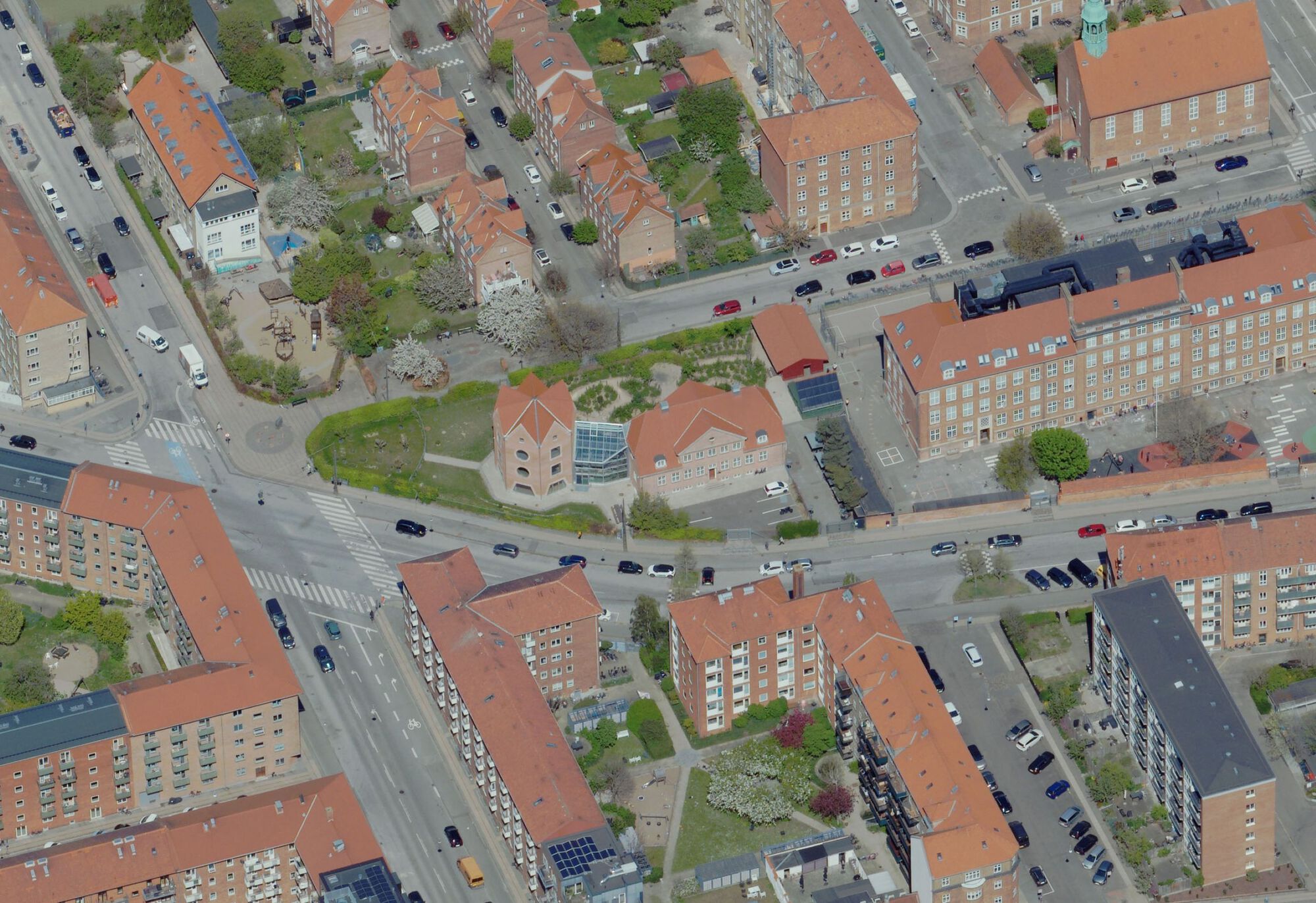Oblique aerial view of a city block of red-roofed brick buildings, streets with parked cars and a green courtyard in spring