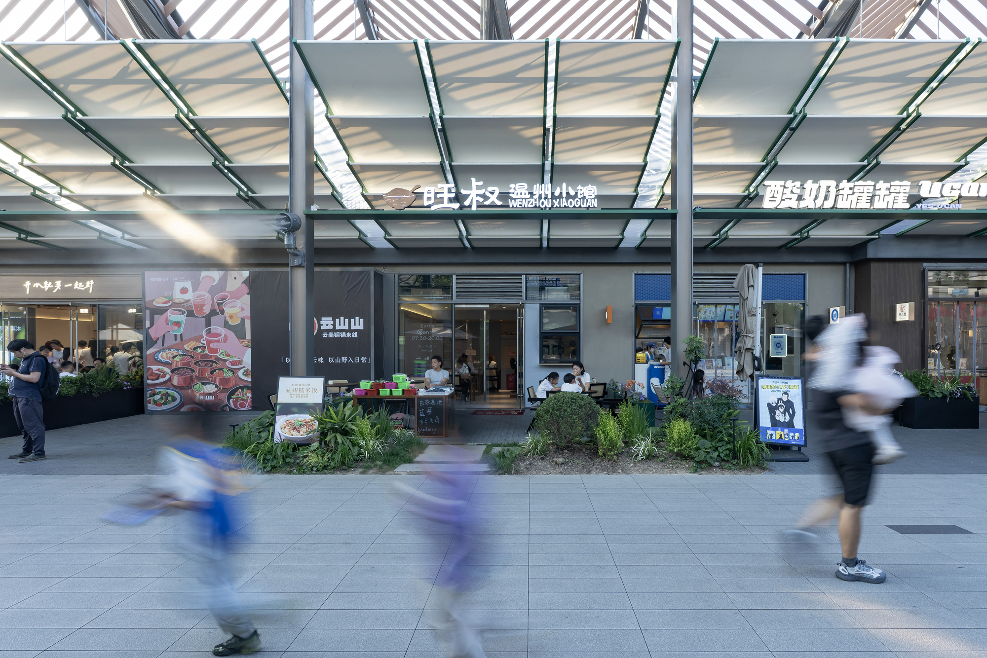 Shopfront arcade beneath a slatted canopy with green steel frames, blurred shoppers crossing the paved plaza in the foreground