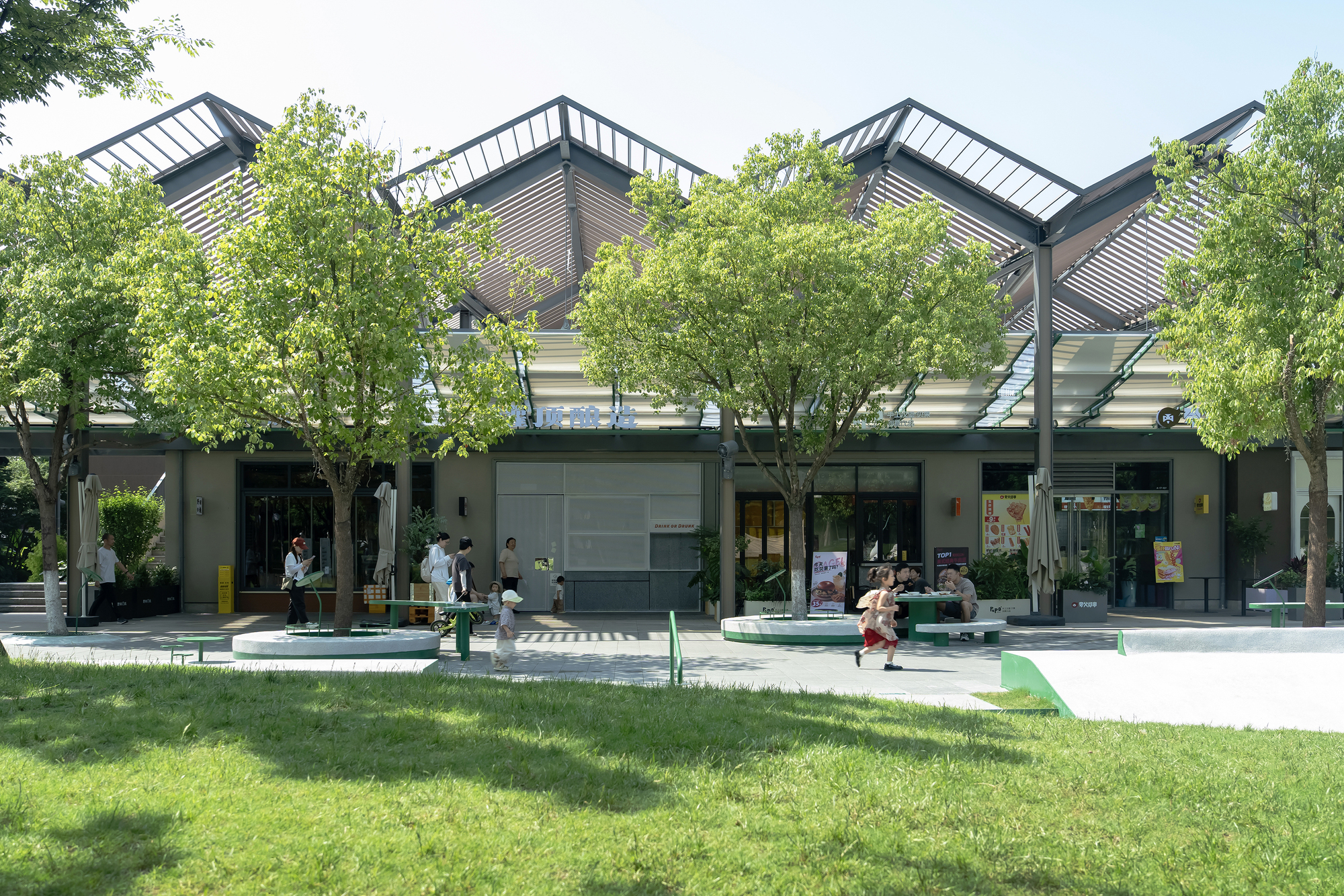 Row of sawtooth louvered roofs behind young trees and a lawn, with a child running across the plaza in sunlight