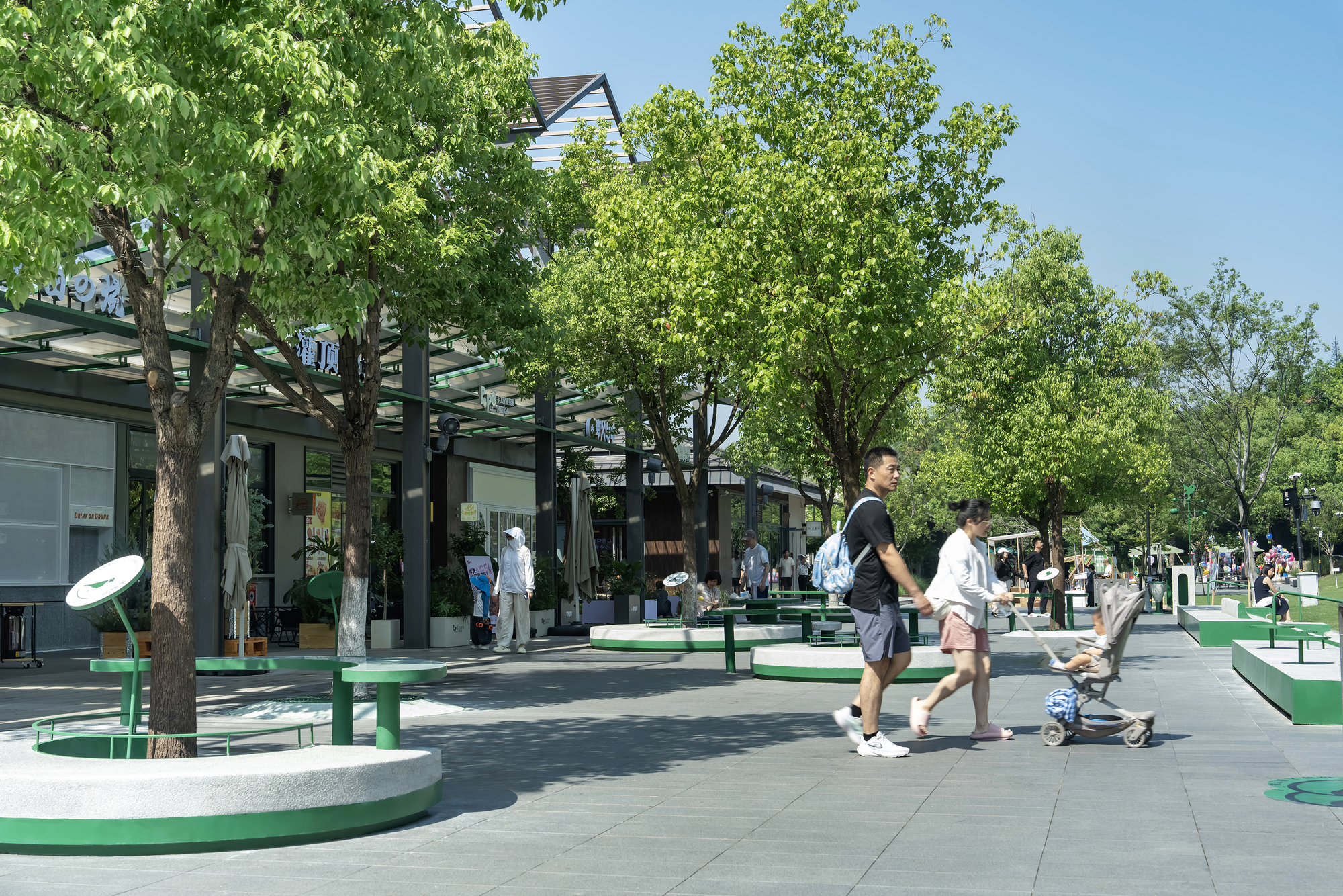 Pedestrian plaza with round green-edged tree benches, a couple pushing a stroller past trees under a clear blue sky