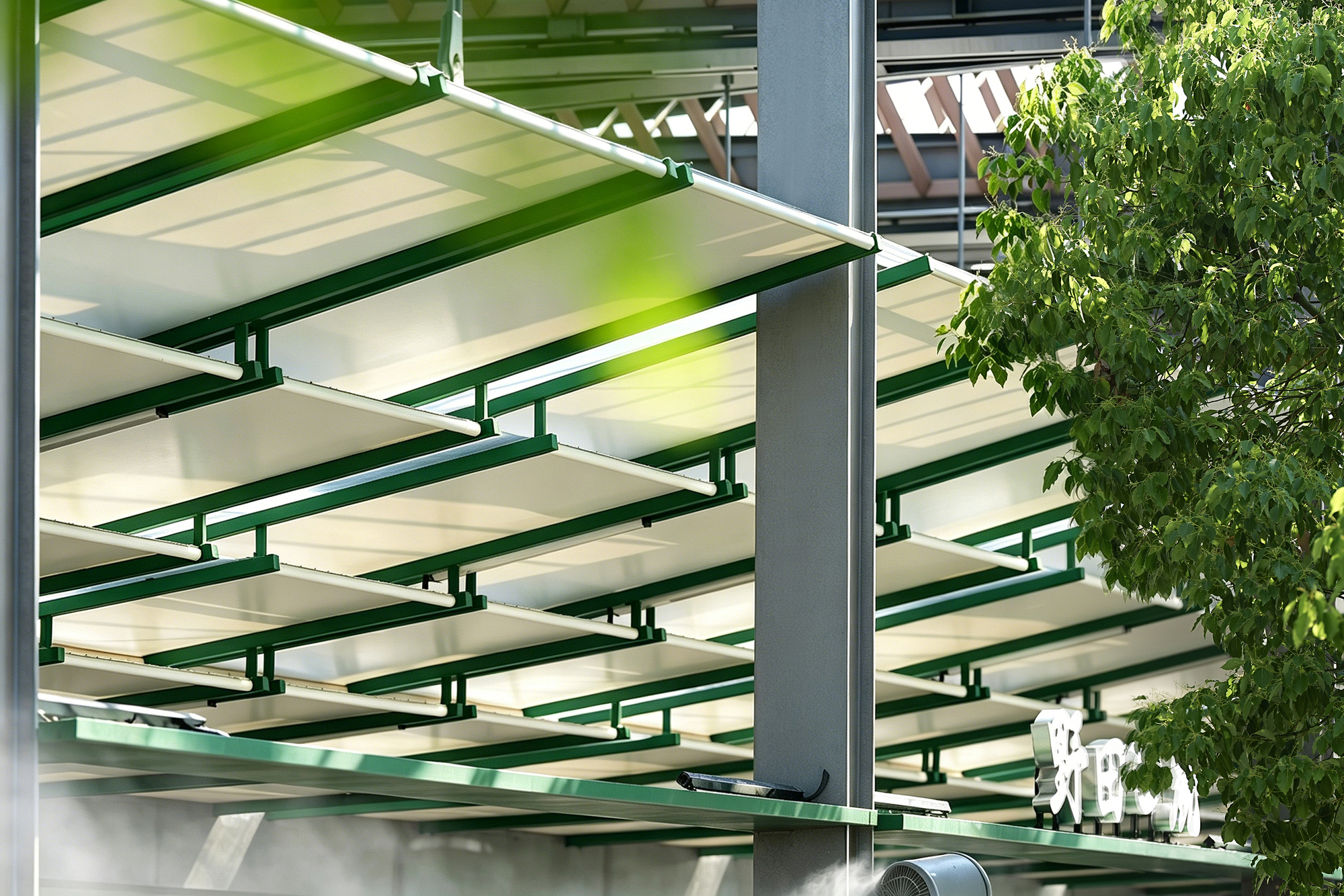 Close-up of the canopy's white panels held by green steel brackets and a grey column, with leaves in sunlight
