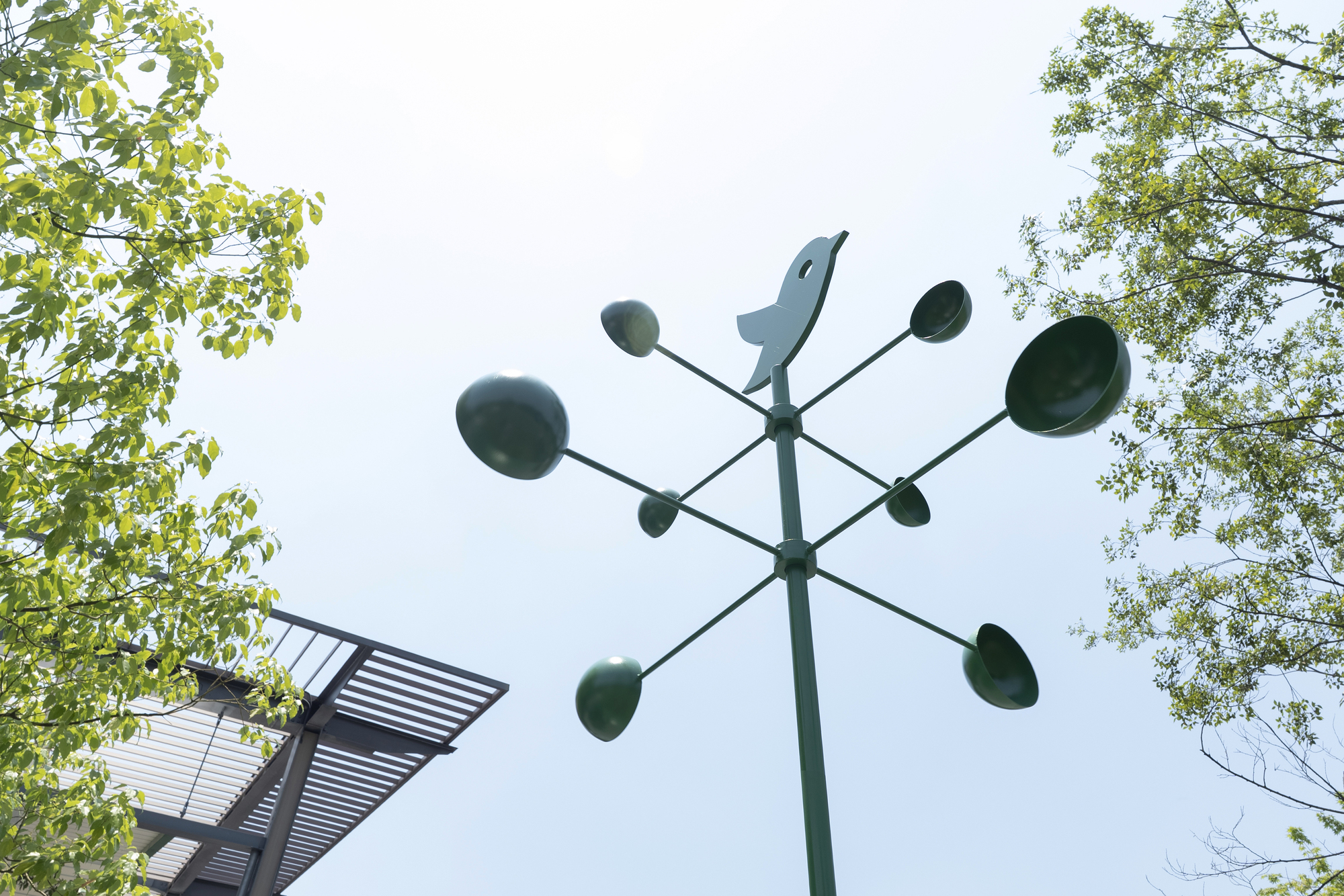 Upward view of a green weathervane with a bird silhouette and cup-shaped arms, framed by leafy branches and a louvered canopy edge