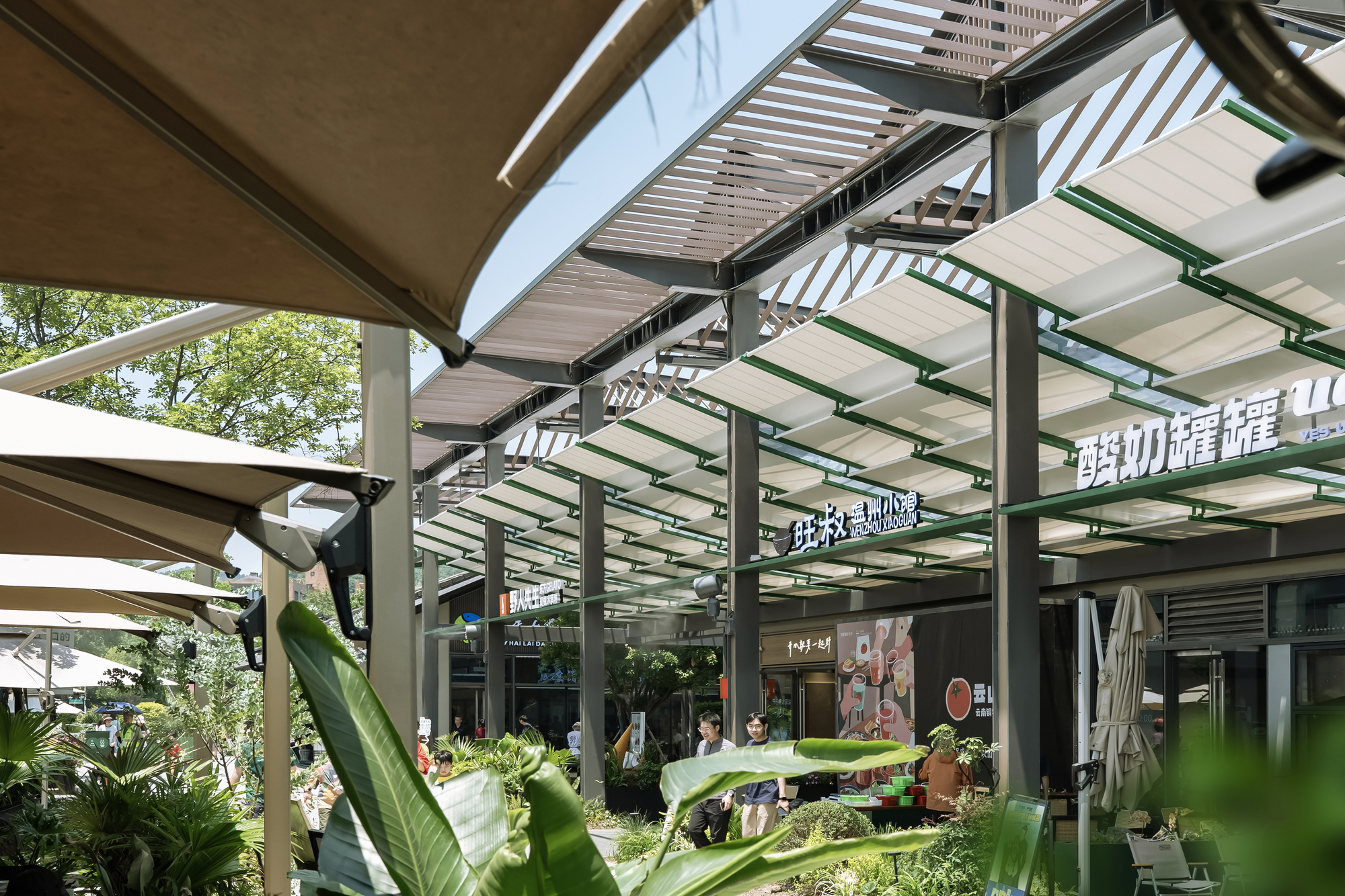 Covered shopping walkway with steel columns, green-trimmed fabric awnings and louvered canopy, seen past banana leaves with diners at the storefronts