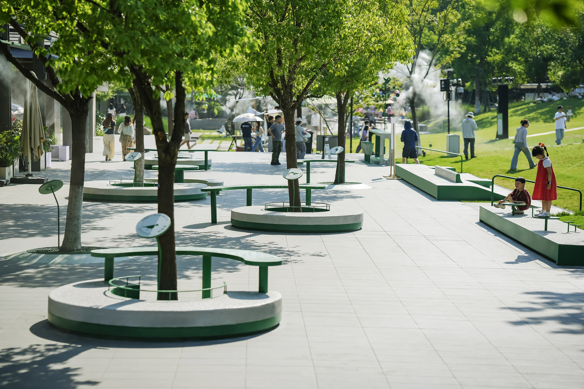 Circular stone planters with green steel benches around young trees, with children and walkers on a sunlit paved plaza beside a lawn