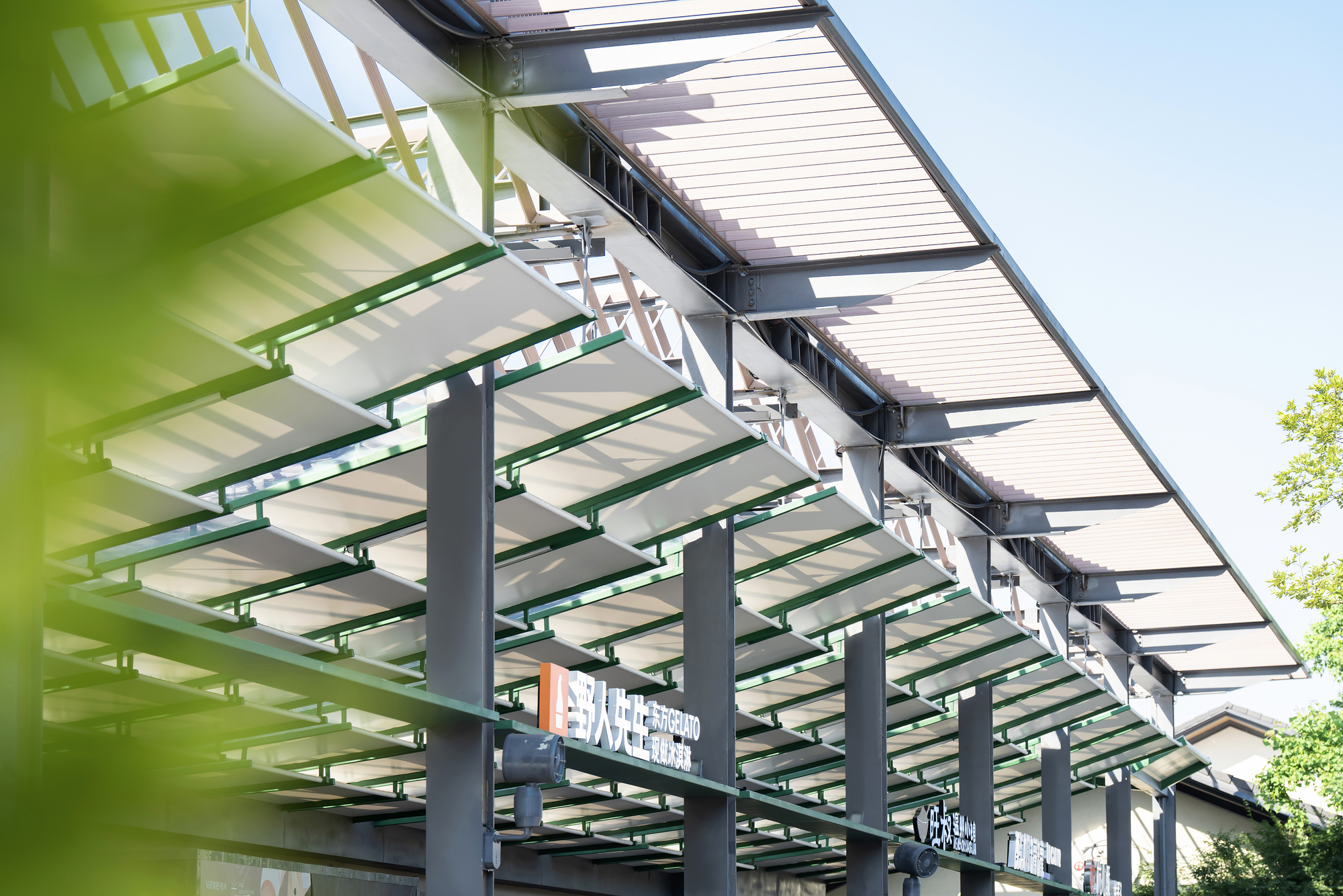 Looking up at the sloped canopy of white louvers on green steel rails, supported by grey columns against a clear blue sky