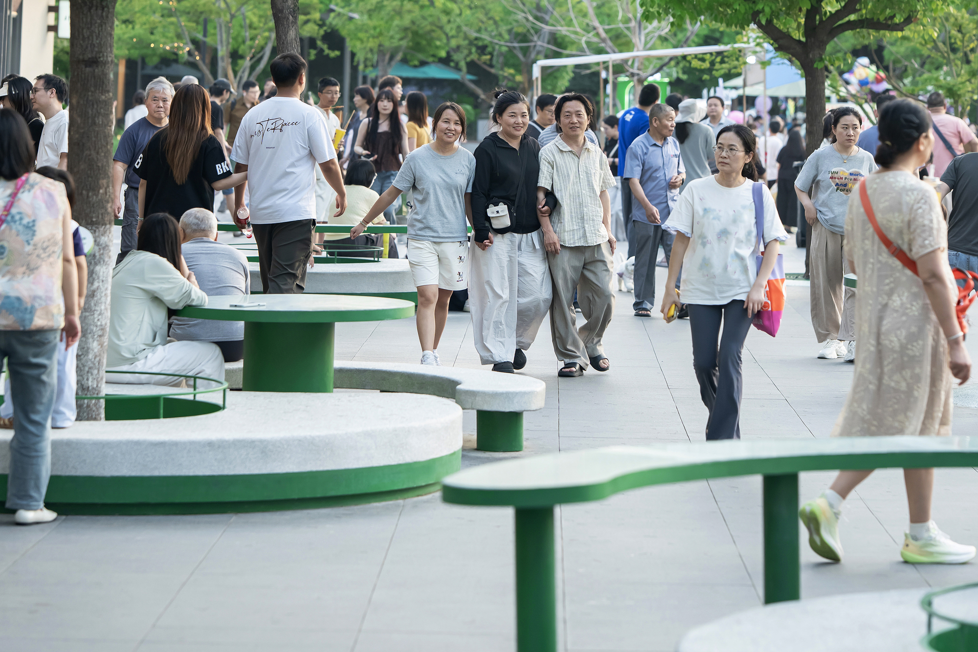 A vibrant public pedestrian promenade featuring custom-designed curved concrete benches and vivid green integrated tables. Large canopy trees provide natural sh
