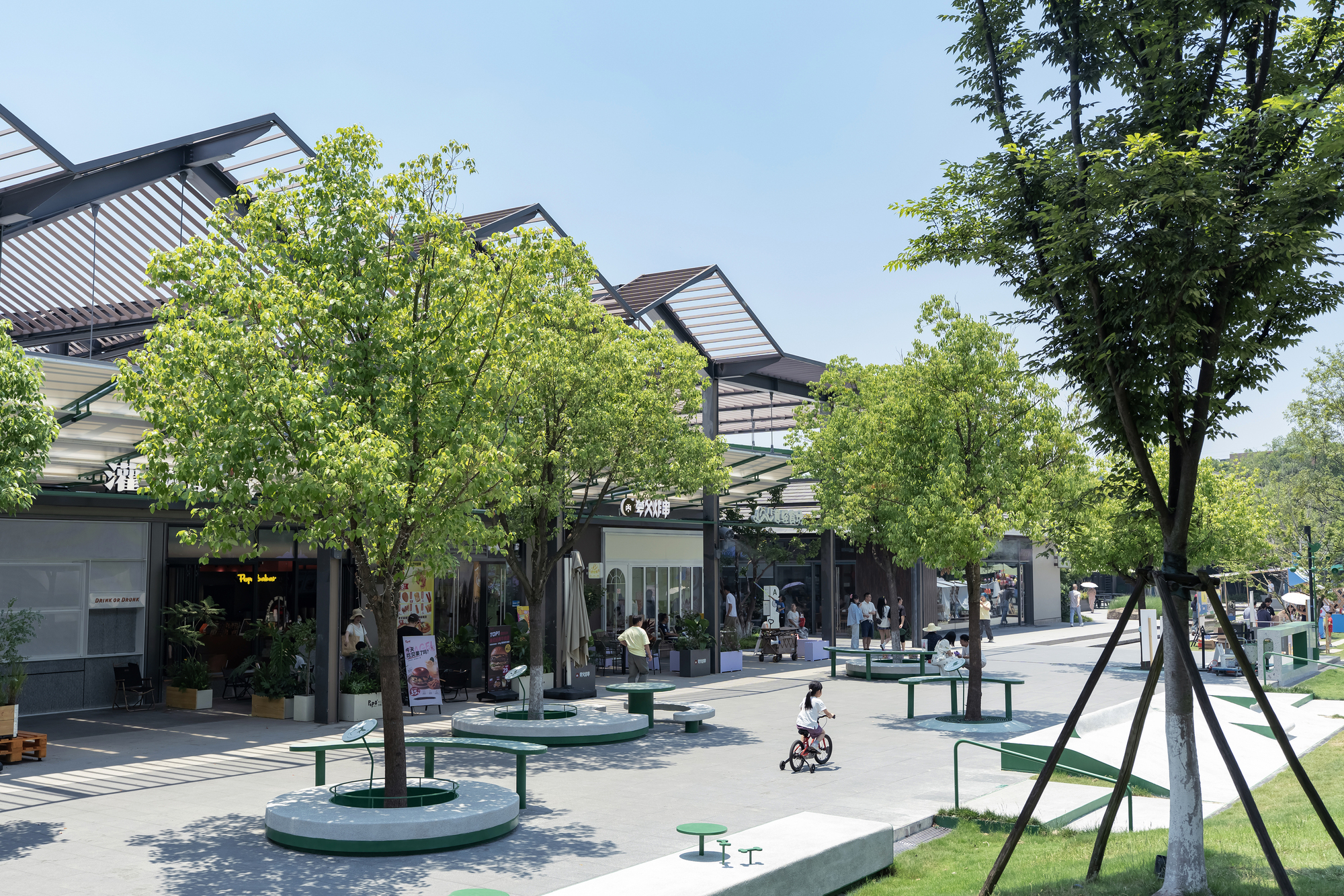 This daytime photograph captures a vibrant pedestrian commercial plaza lined with low-rise retail storefronts beneath an expressive, multi-gabled slatted steel